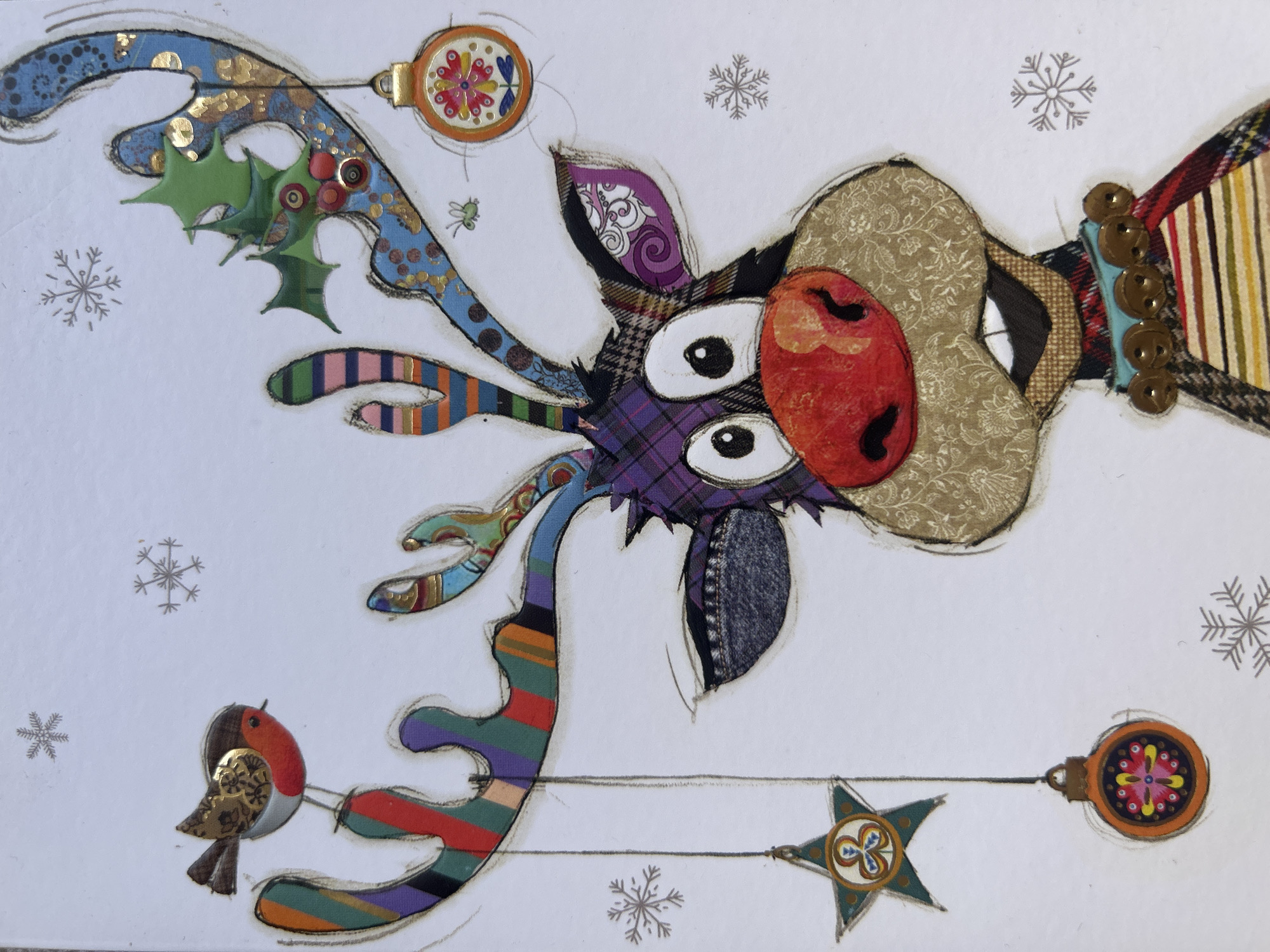 The image depicts a stylized reindeer crafted from various fabric scraps and paper cutouts, arranged against a white background. The reindeer has a large, red nose and mismatched eyes, with antlers decorated with holly, an ornament, and a bird. The background features faint snowflake patterns. Two ornaments and a star are suspended by thin lines below the reindeer. The style suggests a handmade Christmas card or decoration. There is no information in the image to identify the location as Câmara de Lobos, Portugal.