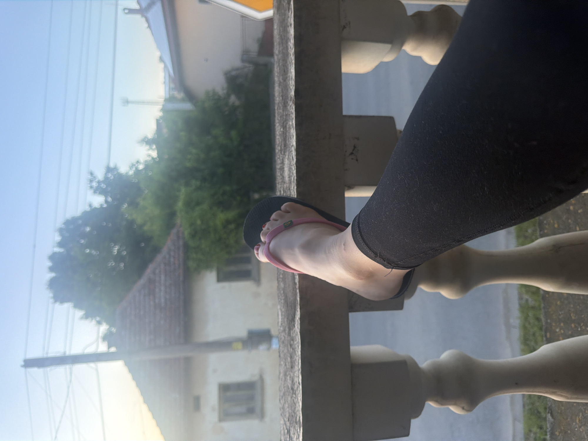 The image, rotated 90 degrees clockwise, presents a close-up view of a person's lower leg and foot resting on a balcony railing, with a residential scene in the background. The foreground prominently features a person's right lower leg, dressed in black leggings, and their foot, adorned with a black flip-flop that has a bright pink strap. The toenails are painted a vibrant pink or red. A small, circular emblem with green and yellow colors is visible on the pink strap. The foot rests casually on a textured, light-colored concrete or stone railing, which features decorative, bulbous balusters extending downwards.
Beyond the railing, the background unfolds into a peaceful residential area under a clear, light blue sky. The soft, diffused lighting suggests it is either early morning or late afternoon, as there are no harsh shadows. Several buildings are scattered across the landscape; one prominent house has a distinct, reddish-brown tiled roof, suggesting traditional architecture. Clusters of green trees and bushes are visible, some appearing softly blurred, adding a sense of depth. In the upper left portion of the sky, multiple thin, parallel lines, likely power or telephone cables, stretch across the frame. A small strip of road or ground with grass is visible beneath the distant buildings. The overall atmosphere conveys a sense of relaxation and observation of the quiet surroundings. No discernible text is visible within the image beyond the small emblem.
