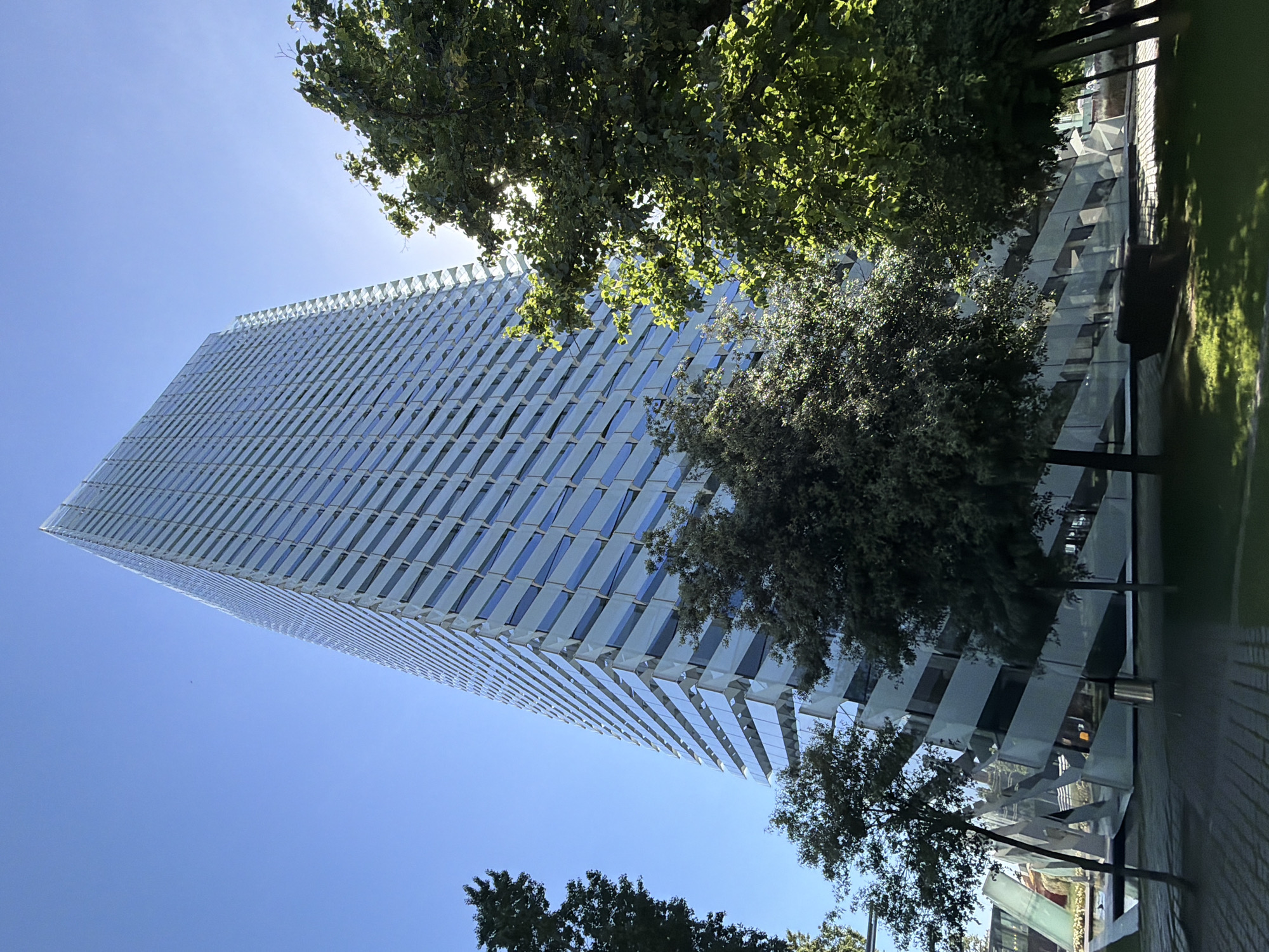 A tall, modern building with a reflective facade is seen from a low-angle, upward-looking perspective in L'Hospitalet de Llobregat, Spain. The building's exterior features a pattern of horizontal, rectangular elements that alternate between a light-colored material and glass windows. Blue sky is visible between the building's top and the surrounding trees. Green trees are located on either side of the building in the image. Pavement is visible on the right side of the image, as is grass. A few lower buildings are visible in the bottom right of the image.
