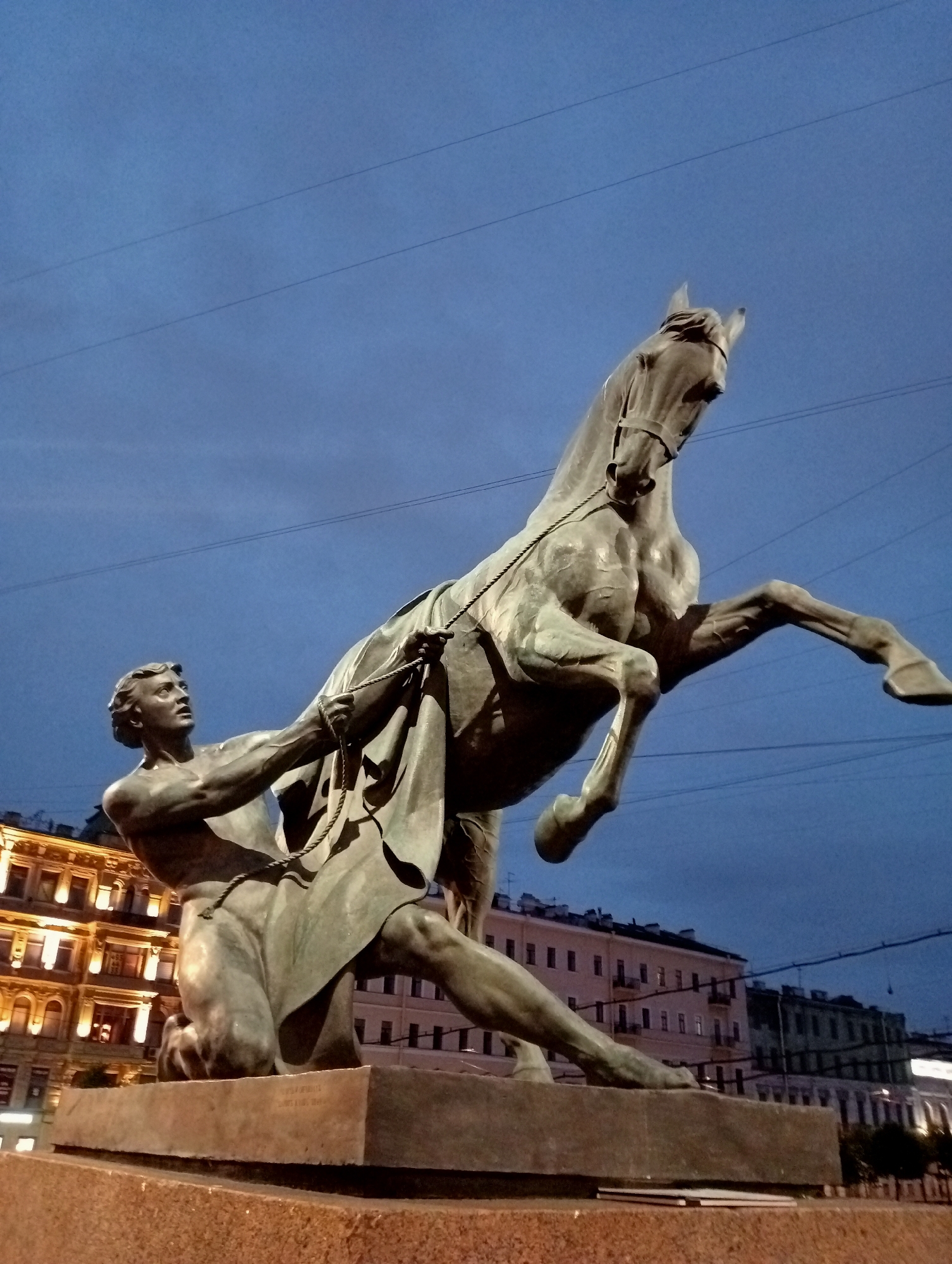 The image depicts a bronze sculpture of a man subduing a rearing horse, set against a twilight sky with visible power lines. The sculpture is positioned in front of buildings that are lit with warm light. The man, depicted with muscular physique, is kneeling and firmly grasping the reins of the horse, which is rearing on its hind legs. The overall scene captures a moment of struggle and control. This sculpture is the Bronze Horseman (Russian: Медный всадник, Mednyy vsadnik), officially known as the Monument to Peter the Great, located in Senate Square in Saint Petersburg, Russia.