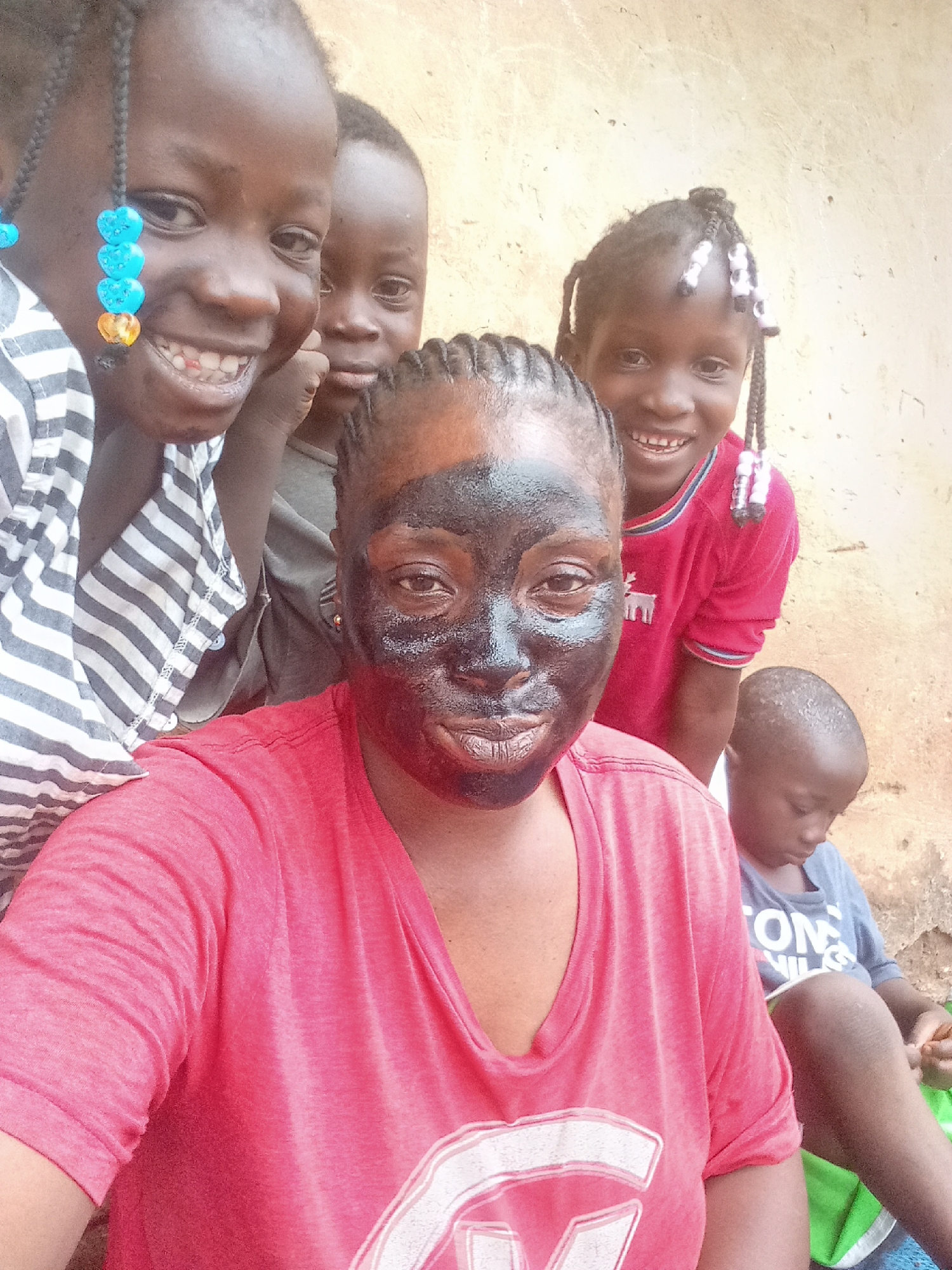 A selfie featuring a woman with a dark facial mask is in the foreground, with four children behind her. The woman has a red shirt on and braided hair. The children are smiling and looking at the camera. The child closest to the woman has a striped shirt and beads in their braids. Another child has a red shirt, and there is a third child in a gray shirt. A small child sitting on the ground to the right has a gray shirt with white letters. They appear to be outside, near a light-colored wall. The location is presumed to be Akwanga, Nigeria.