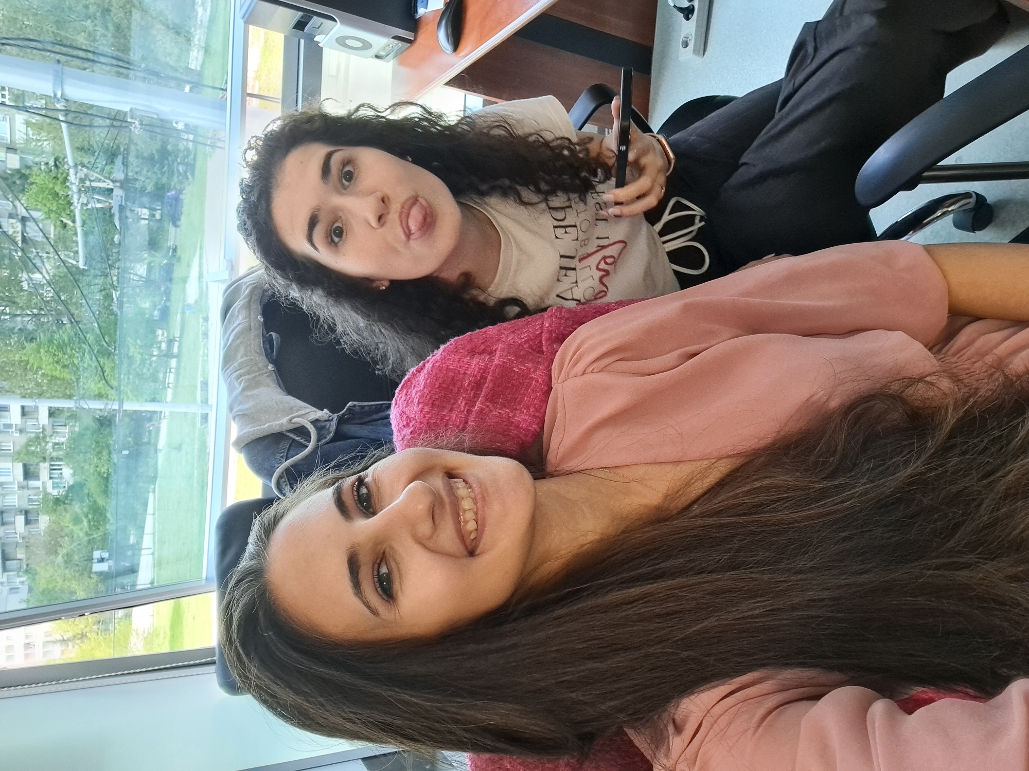 The image, originally rotated 90 degrees counter-clockwise, portrays two young women in an indoor setting, possibly an office or study room, in what appears to be a casual selfie or photograph.
In the foreground, a woman with long, dark, straight hair is seen from the chest up, looking directly at the camera with a wide, cheerful smile. She is wearing a pink long-sleeved top and a thin necklace. Her hair cascades over her shoulders and part of the frame.
Behind her, another woman with dark, curly hair is visible from the waist up. She is making a playful expression, sticking her tongue out and looking at the camera with wide eyes. She wears a white short-sleeved t-shirt with the mirrored text "LA VIE EST BELLE" (Life is Beautiful), with "est belle" highlighted in red. She holds a black smartphone in her right hand and has a gold-colored bracelet on her left wrist. She is seated in a black office chair, with her dark pants visible.
The background features a large window offering a view of an outdoor scene, including green trees, grass, and residential buildings under natural daylight, suggesting it is daytime. A desk with computer equipment and other items is partially visible behind the women. The overall mood is lighthearted and joyful.