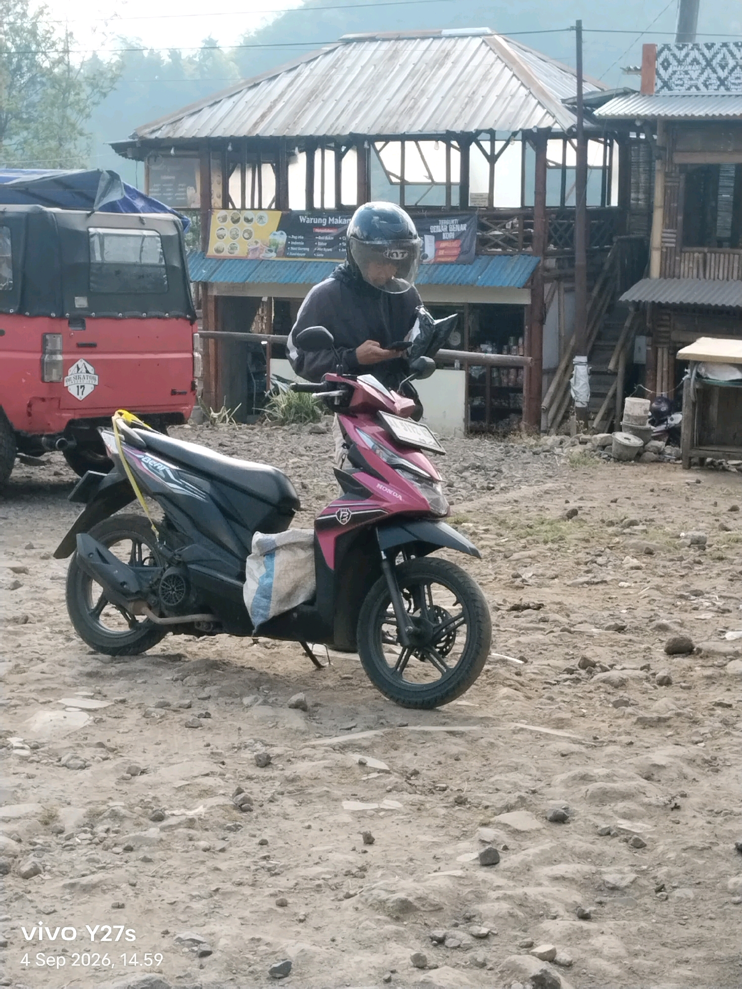 A person wearing a dark jacket and a black helmet is sitting on a pink and black motorcycle. The person is looking down at a phone in their hands. The motorcycle is parked on a dirt road in front of a rustic building with a tin roof. To the left of the motorcycle is the rear of a red vehicle. The background shows a hazy hillside with trees. The scene appears to be in a rural or semi-rural area. The timestamp indicates the photo was taken on September 4, 2026, at 14:59 in Sigedang, Indonesia.