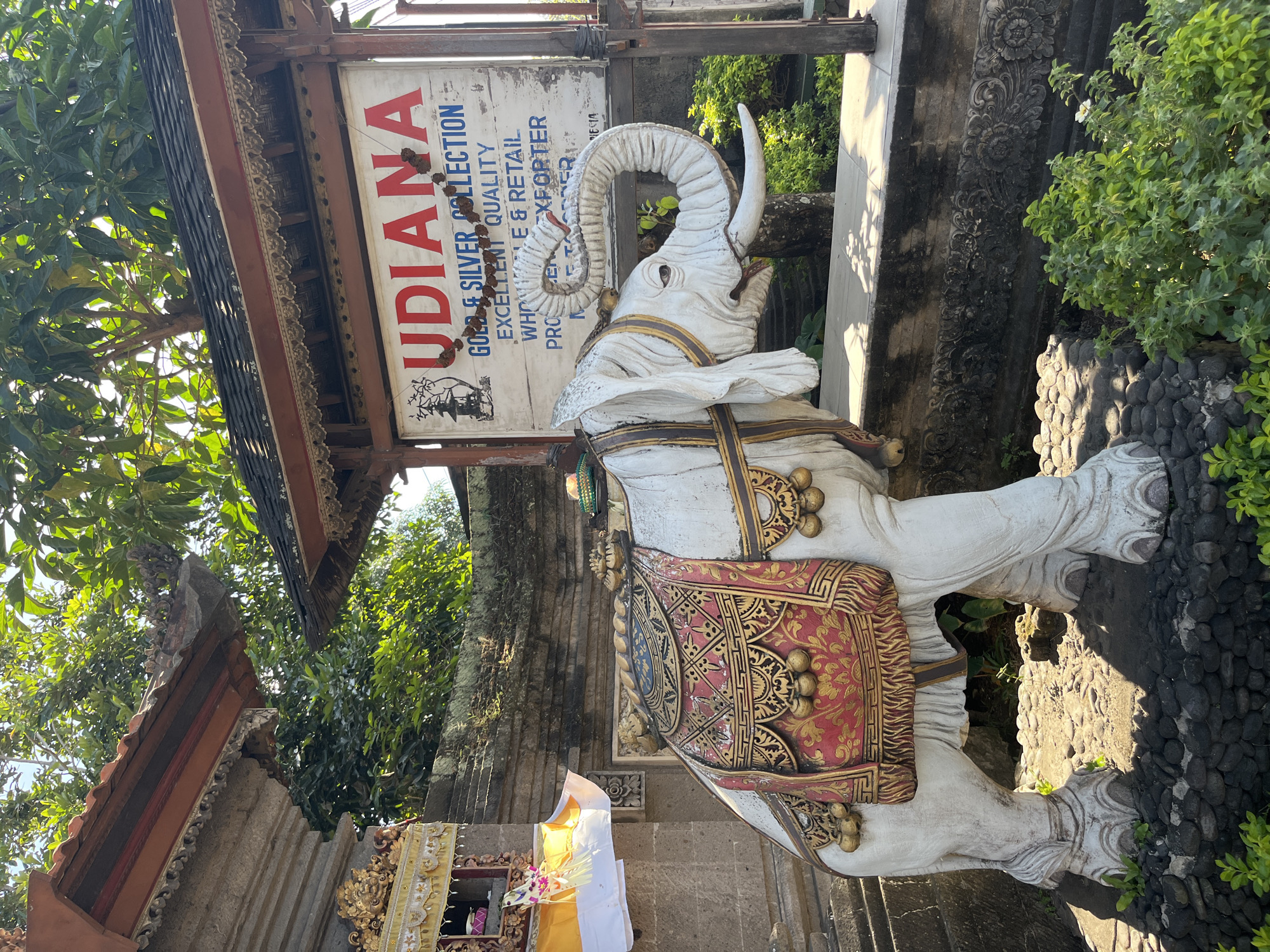 A statue of a white elephant adorned with a decorative saddle stands in front of a building with a sign that reads "UDIANA GOLD & SILVER COLLECTION". The elephant is a realistic depiction, with intricate details in its skin texture and tusks. The saddle is richly decorated with red and gold patterns, and small golden balls hang from its sides. Behind the elephant and to the left, a stone structure with a multi-tiered roof and a small shrine adorned with white and yellow fabric is visible. To the right of the elephant, the sign is mounted on a wooden frame, and further back, lush green trees and foliage are present. The ground around the elephant is covered with small stones, and a low stone wall with carved patterns is behind it. The scene is bathed in natural sunlight, suggesting it is daytime. The location is identified as Sukawati, Indonesia.