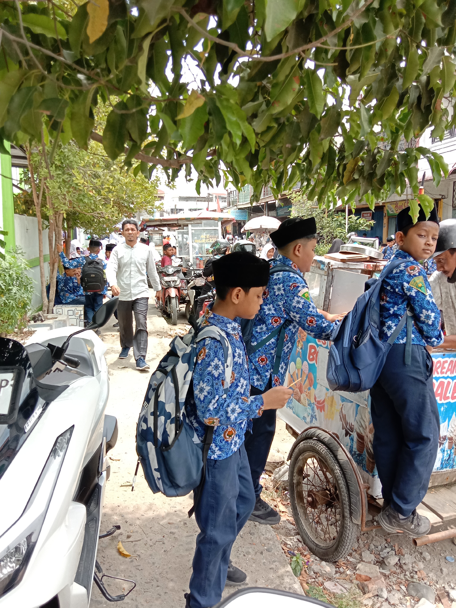 In Lhokseumawe, Indonesia, a group of young boys in school uniforms, consisting of blue patterned shirts and dark pants, are gathered around an ice cream cart. The boys are wearing black peci hats. One boy in the foreground, wearing a blue and white patterned shirt with a camouflage backpack, is holding a stick and appears to be interacting with the cart. To his left, another boy in a similar uniform with a blue backpack is also near the cart. Further back, a man in a light-colored collared shirt and dark pants is walking away from the camera. The environment appears to be a street scene with trees, buildings, and parked motorcycles visible. An ice cream cart with colorful advertisements for ice cream is the central focus of the children's attention.