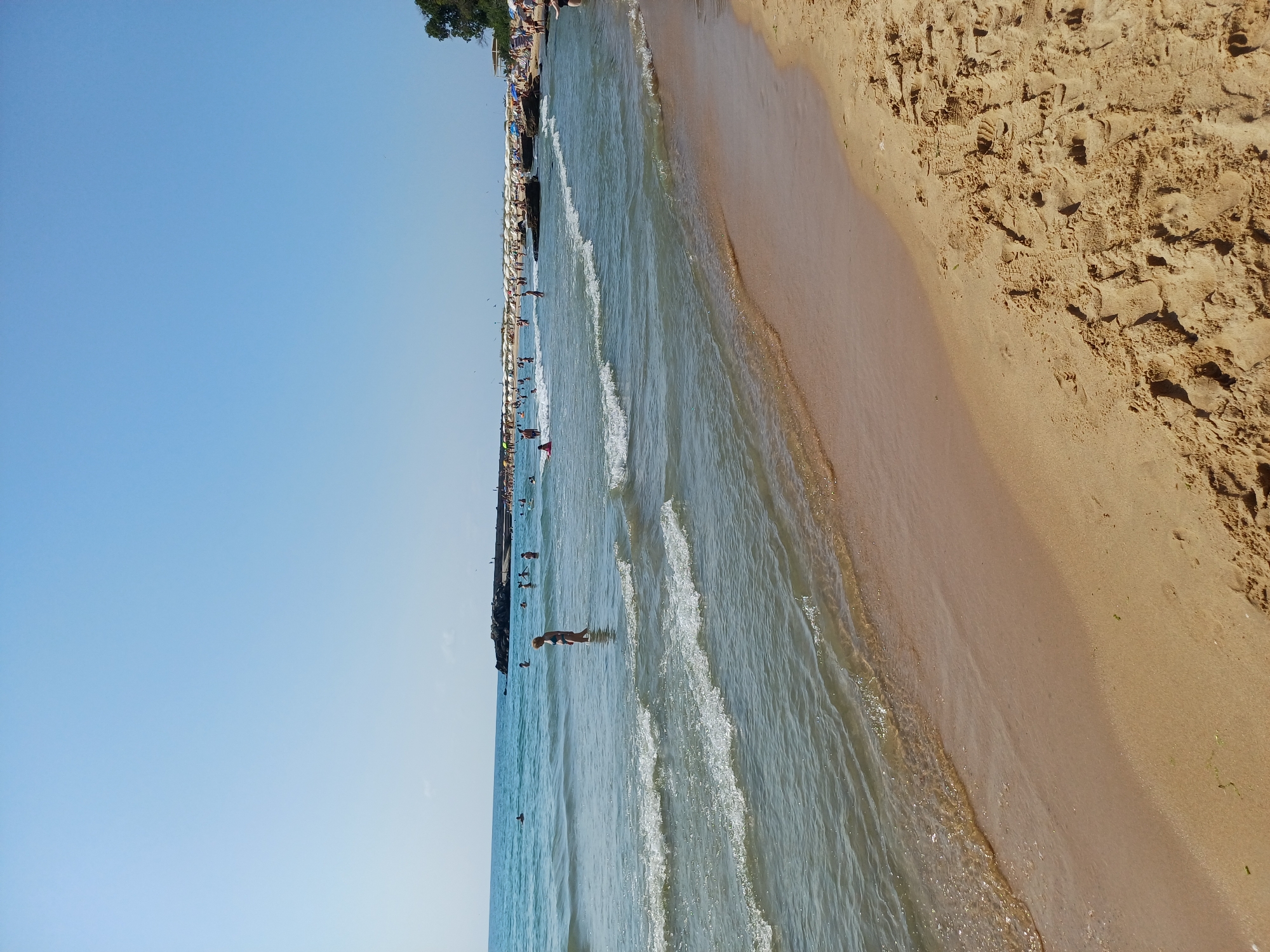 The image depicts a busy beach scene on a clear, sunny day, likely at the Varna City Beach in Varna, Bulgaria. The foreground is dominated by sand with footprints and some scattered seaweed. Gentle waves are lapping at the shore. In the shallow water, several people are visible, some wading and others standing. Further out, the sea is calm, and a breakwater or pier extends into the water on the left side of the frame. A crowd of people are gathered along the waterline and on the beach in the distance, with many beach umbrellas providing shade. Trees are visible on the far right, suggesting the presence of a promenade or park area behind the beach. The overall impression is one of a vibrant summer day at a popular seaside location.