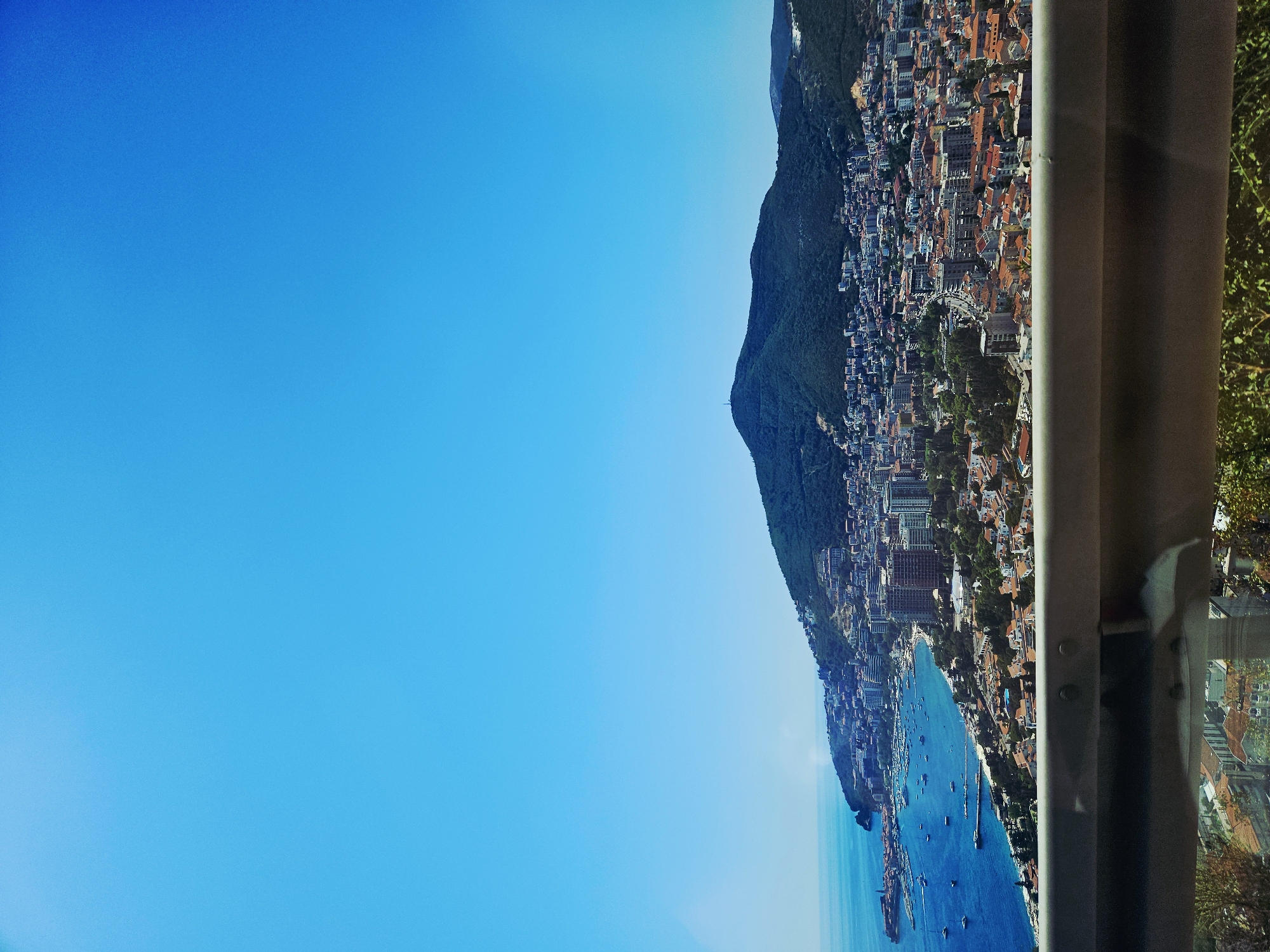 The image captures a panoramic view of a coastal city nestled against a lush green mountainside under a clear blue sky. In the foreground, a metal guardrail runs horizontally across the frame, partially obscuring the view of the city below. Beyond the guardrail, the city sprawls across the slopes, characterized by a dense collection of buildings with red-tiled roofs and modern high-rise structures. A vibrant blue bay, dotted with numerous boats and yachts, forms the city's coastline. The water is calm, reflecting the bright sunlight. To the left, a historic-looking fortified town juts out into the sea. The background is dominated by a large, densely forested mountain that rises majestically behind the city. The scene appears to be taken from an elevated vantage point, possibly a viewpoint or a road winding up the mountain, offering a scenic perspective of the urban landscape and its natural surroundings. The location is identified as Košljun, Montenegro, suggesting a view overlooking the Adriatic coast.