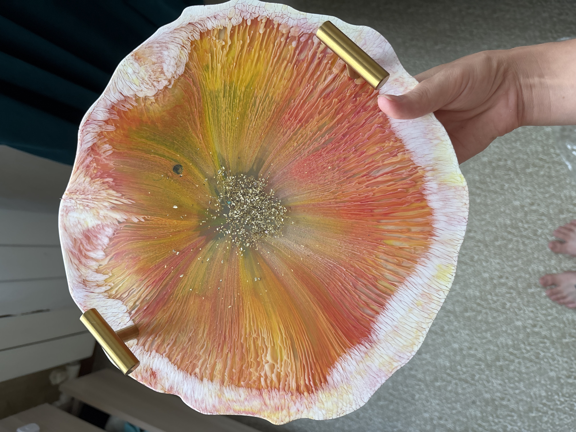 A person's hand holds a round, decorative tray with a colorful, abstract pattern resembling a flower or a splash of liquid. The tray has two brass handles. The background is blurred, showing what appears to be indoor furniture and carpeting. There are no discernible landmarks or clues to indicate a specific location in Naberezhnye Chelny, Russia. The scene appears to be an indoor setting, possibly a home.