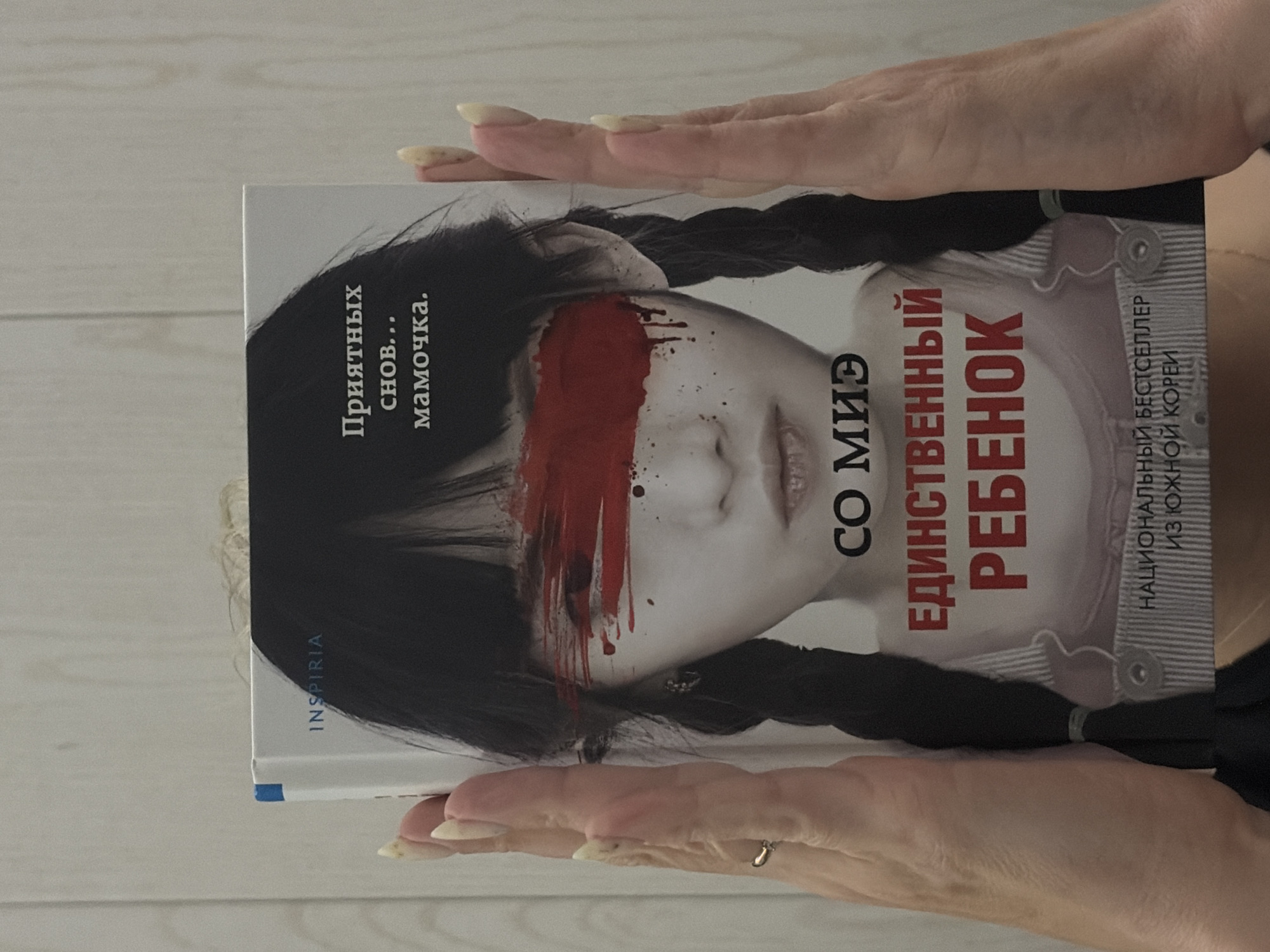 The image shows a book held by two hands. The book cover features a black and white portrait of a young girl with dark braids. A red streak covers her eyes. The title, written in Russian, includes "Единственный Ребенок", meaning "Only Child". The text also includes the author's name, "CO МИЭ" (So Mie), and the phrase "Приятных снов... мамочка" (Sweet dreams... Mommy). The spine displays "INSPIRIA". On the right side, it says "НАЦИОНАЛЬНЫЙ БЕСТСЕЛЛЕР ИЗ ЮЖНОЙ КОРЕЙ" (National Bestseller from South Korea). The background is a light wood grain. There is no evidence to suggest this image was taken in the city of Chaikovsky, Russia.