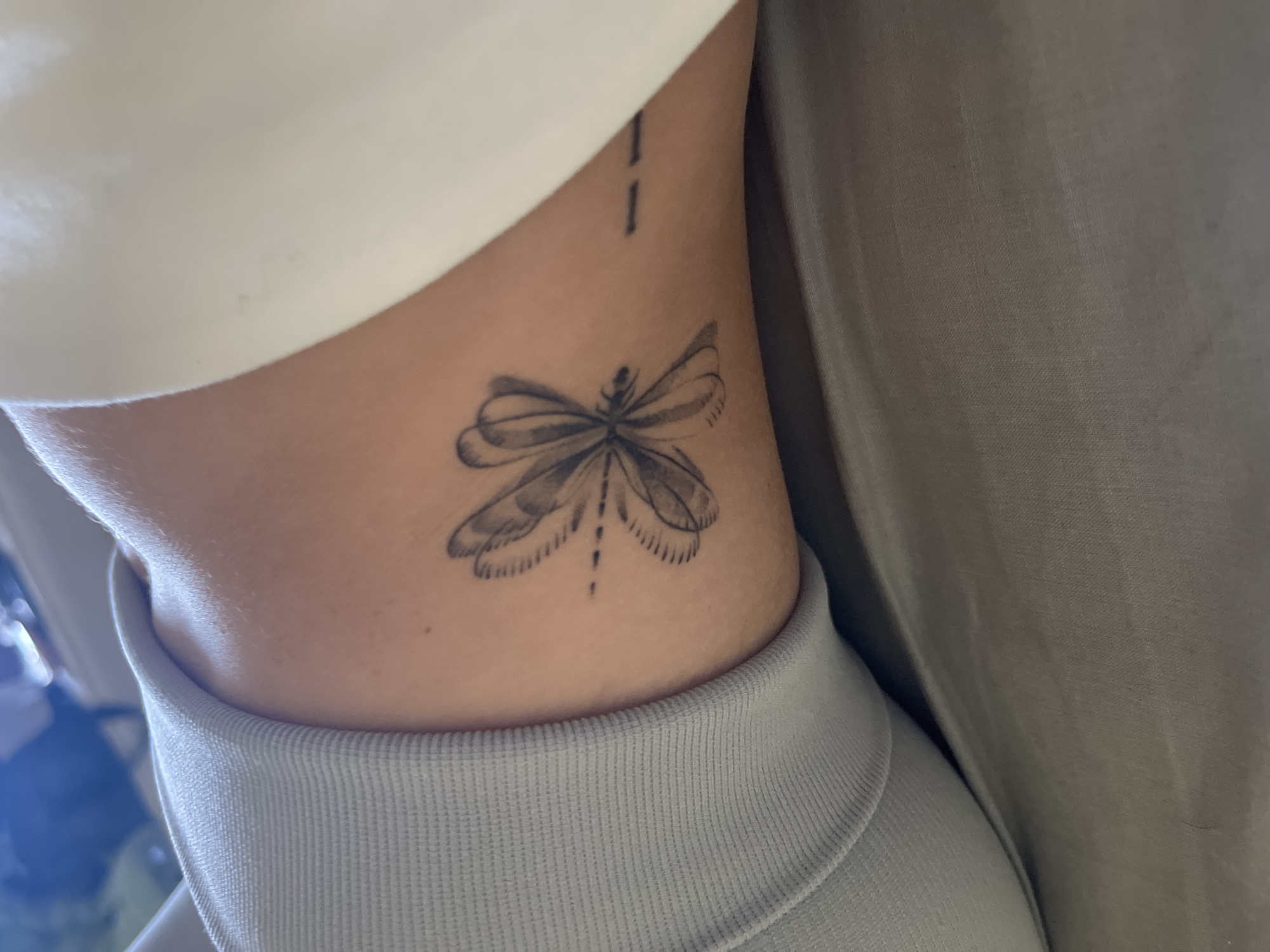 An extreme close-up shot shows a tattoo of a dragonfly on a person's arm, which appears to be a pale complexion. The dragonfly is outlined in black and shaded with grey ink. Its wings are depicted with a transparent, layered effect, and its body is simplified. To the right of the dragonfly, a series of short, horizontal black lines are tattooed, appearing as a dash and a sequence of three dashes. The arm is bent, and a grey, ribbed fabric, possibly clothing, is visible on the left side of the frame, creating a soft focus background. The lighting suggests an indoor setting, potentially with natural light illuminating the scene from above and the right.