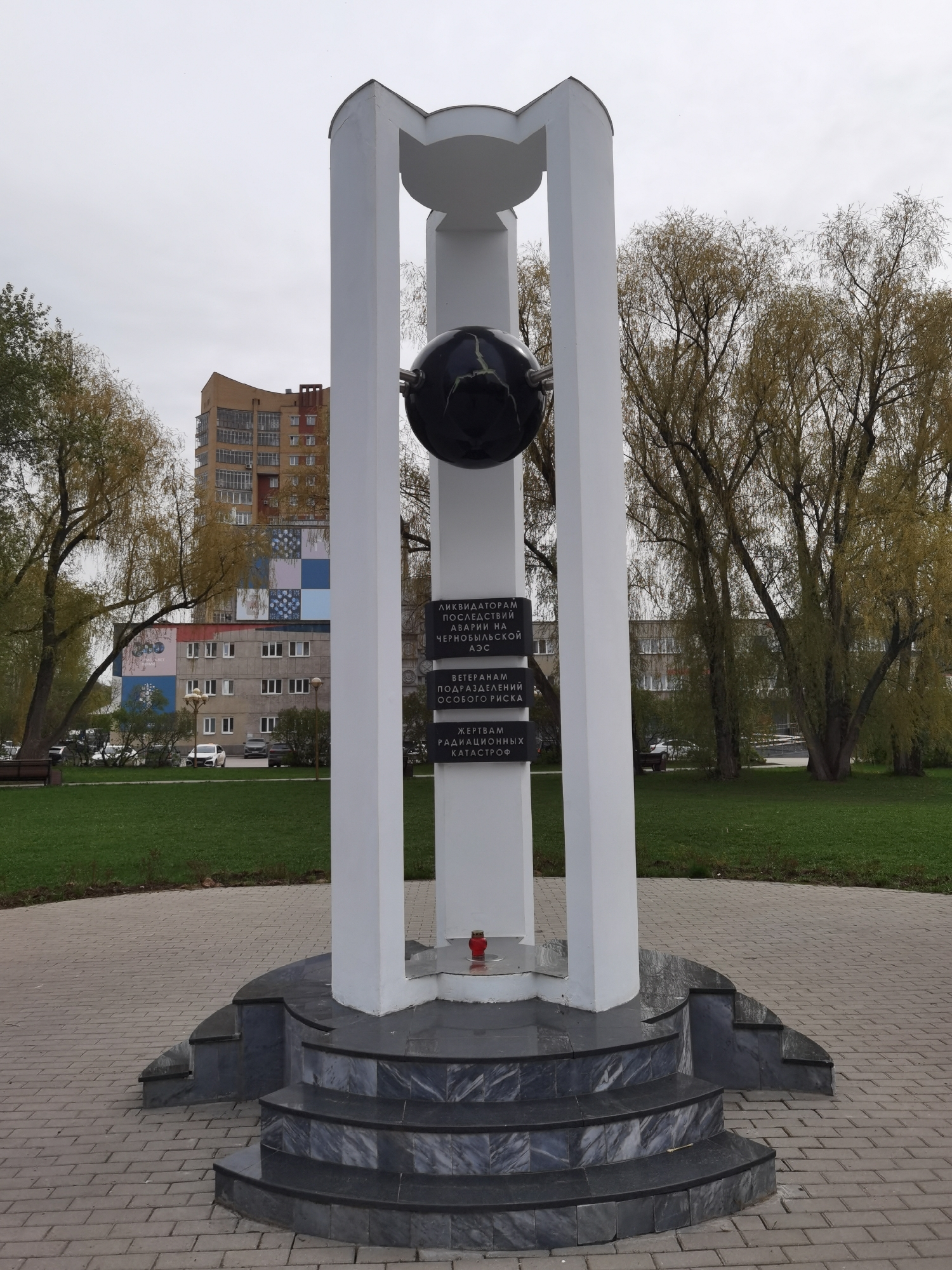 The image displays a commemorative monument located in an urban park or square in Perm, Russia, under an overcast sky. The central subject is a tall, white structure with two arched pillars framing a central column. Suspended within this framework is a dark, spherical object, possibly a globe or an abstract representation.
Three dark plaques with white Cyrillic text are affixed to the central column. The top plaque reads: "ЛИКВИДАТОРАМ ПОСЛЕДСТВИЙ АВАРИИ НА ЧЕРНОБЫЛЬСКОЙ АЭС" (To the liquidators of the consequences of the Chernobyl accident). The middle plaque states: "ВЕТЕРАНАМ ПОДРАЗДЕЛЕНИЙ ОСОБОГО РИСКА" (To the veterans of special risk units). The bottom plaque says: "ЖЕРТВАМ РАДИАЦИОННЫХ КАТАСТРОФ" (To the victims of radiation catastrophes).
The monument stands on a multi-tiered base of dark, polished marble or stone, featuring curved steps. A single red candle in a glass holder is placed on the base, signifying remembrance.
The setting is an outdoor area with a paved brick pathway leading to the monument, surrounded by green grass. Trees, possibly willows with sparse, light green foliage, flank the monument, suggesting early spring or late autumn. In the background, several multi-story residential buildings are visible, one with a distinctive checkered facade of colored squares. No people are present in the scene. The overall impression is solemn and commemorative, dedicated to those affected by radiation catastrophes.