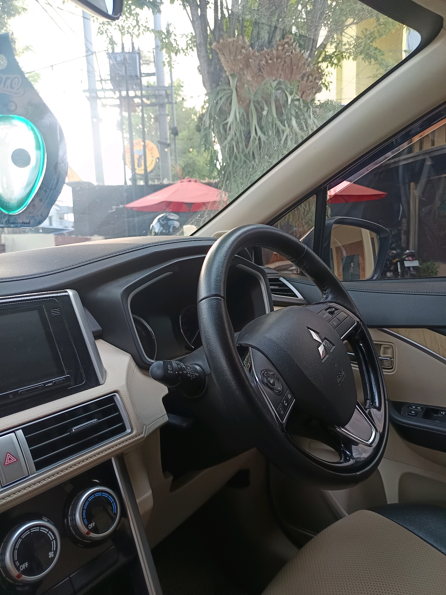 The image shows the interior of a car, likely parked outdoors in Tamansari, Indonesia, as indicated by the presence of tropical foliage and a bright, sunny atmosphere visible through the windshield. The driver's seat is unoccupied. Prominently featured is the black steering wheel with the Mitsubishi logo at its center, and control buttons on its spokes. The dashboard displays a black infotainment screen, air conditioning vents, and climate control knobs. In the background, through the windshield and side windows, are glimpses of the external environment: a tree with epiphytic plants, power lines, a red umbrella, and a building with yellow siding. The interior of the car is primarily composed of beige and black upholstery and trim.