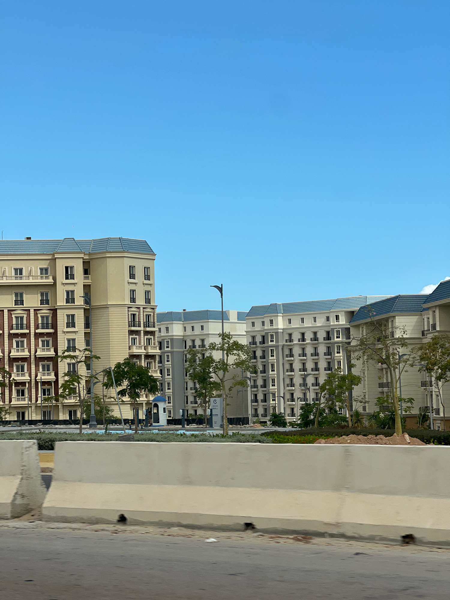 The image captures a section of a modern residential development in El Alamein, Egypt, under a clear blue sky. In the foreground, a concrete barrier separates a road from a landscaped area. Behind the barrier, several large apartment buildings with light-colored facades and blue tiled roofs are visible. The buildings feature numerous balconies, windows, and architectural details. Trees and greenery are interspersed between the buildings, suggesting an urban or suburban setting. The overall scene depicts new construction or recently completed development.
