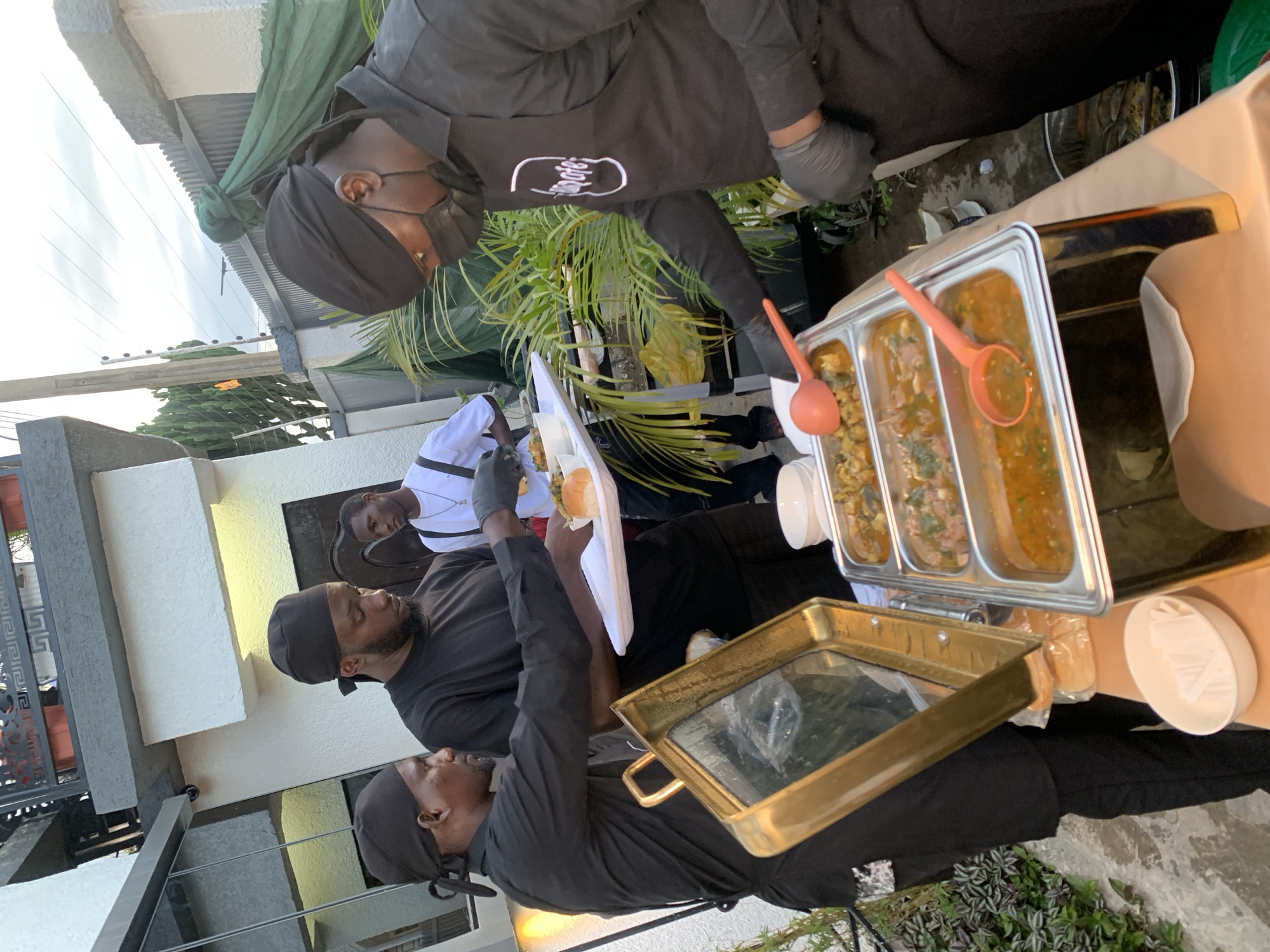 In Gura Top, Nigeria, at an outdoor event, three men are shown interacting around a buffet table. Two men, dressed in black head coverings and black shirts, are serving food from stainless steel chafing dishes. The man on the left, wearing a black head covering, is serving food onto a white plate held by a person out of frame. The man in the center, also in a black head covering and black shirt, is looking down at the food. The third man, wearing a white shirt and black suspenders, stands to the right, observing. He has a black face mask on. The buffet table holds three chafing dishes filled with various stews and a separate dish with bread rolls. A golden chafing dish lid is propped open. Behind the buffet, potted plants and a green draped fabric are visible, suggesting an outdoor gathering. The background shows a building with white walls and reflective windows. The overall scene depicts a catering service in progress.
