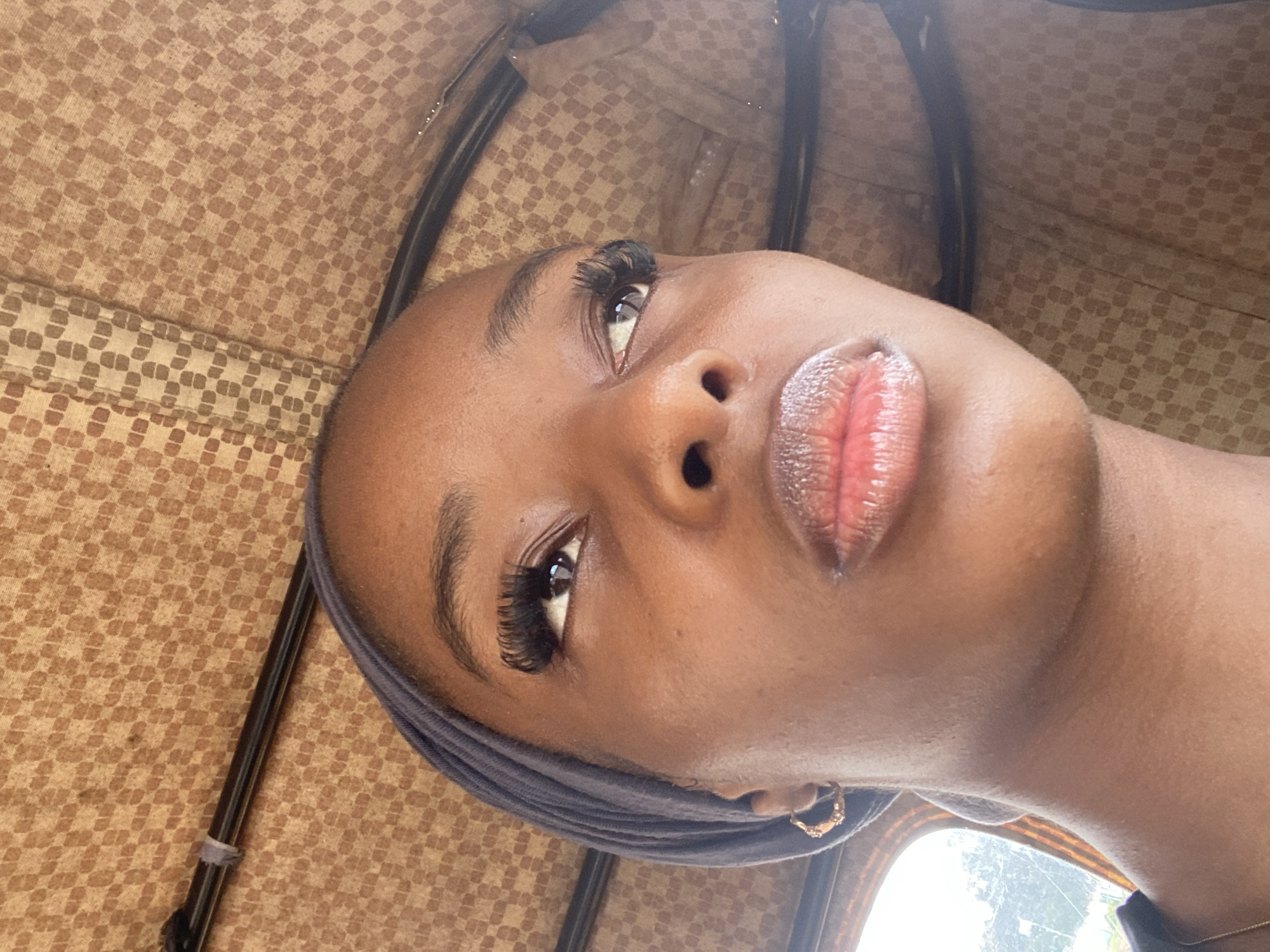 The image shows a close-up of a young Black woman's face, tilted upwards and to the right. She is wearing a dark grey headscarf that covers her hair and drapes around her neck. She has long, dramatic false eyelashes and her lips are slightly parted, revealing a glossy finish from lip gloss or balm. A small, gold hoop earring is visible on her lower earlobe.
The background is a textured, patterned fabric in shades of brown and beige, resembling upholstery or a woven material. A dark, thin pole runs vertically along the left side of the image, and another similar pole or support is visible horizontally at the top. A hint of a window with bright light filtering through suggests an interior setting, possibly a vehicle.
The lighting is bright and somewhat diffuse, suggesting it could be daytime, but the specific time of day is not clear. There are no visible text elements in the image. The overall impression is a portrait with a focus on the subject's facial features and makeup, set against a subtly textured backdrop. The location context of Jos, Nigeria, doesn't provide specific visual cues within the image itself but may inform the interpretation of the subject's appearance or environment.