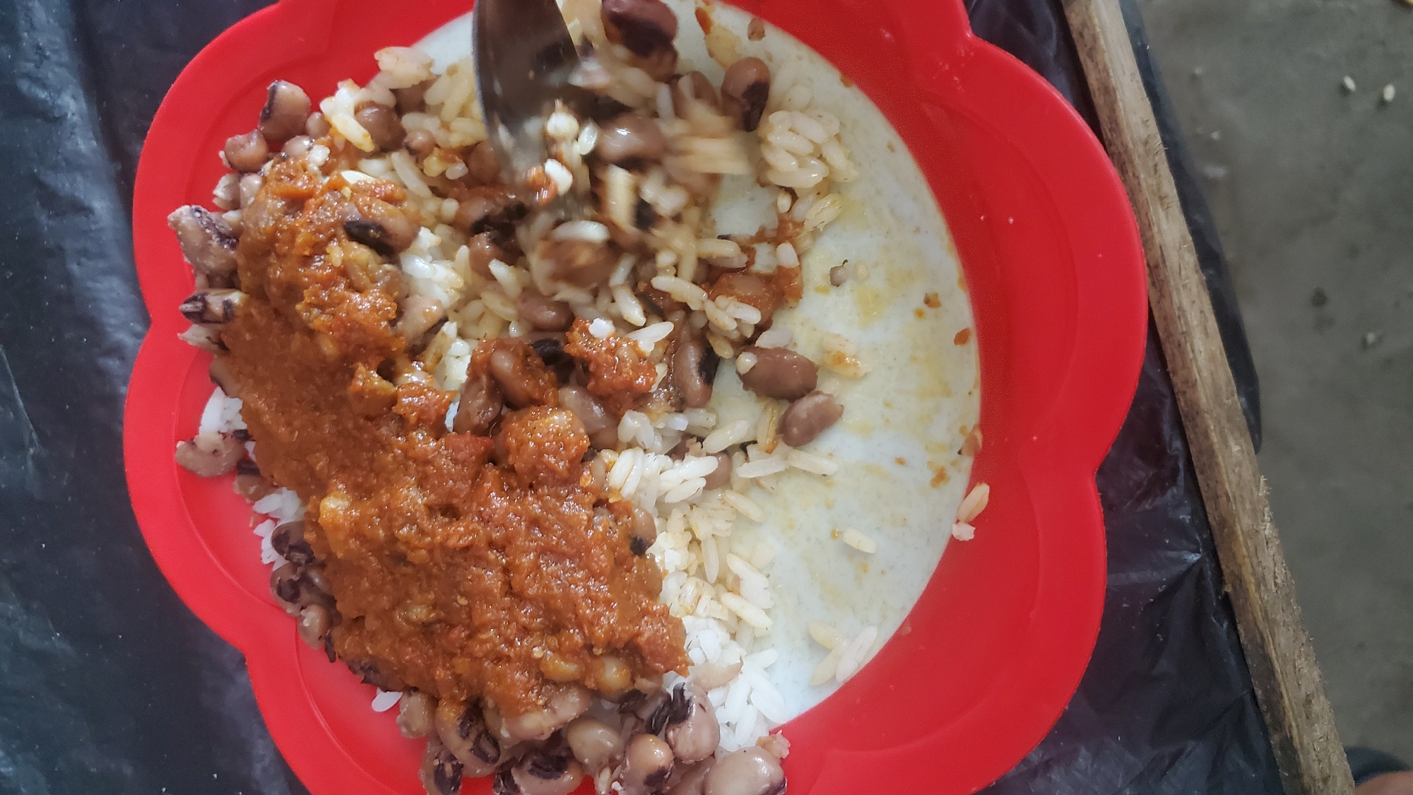 In Jalingo, Nigeria, a red, scalloped plastic plate contains a meal of white rice and black-eyed peas. The peas are mixed with the rice, and a reddish-brown sauce, appearing to be a stew or relish, is spread over the top. A spoon is visible on the right side of the plate, partially submerged in the rice and peas, suggesting the meal is being eaten. The plate rests on a dark, textured surface, possibly a tablecloth or plastic sheeting, with a wooden edge visible in the foreground. The lighting is uneven, with shadows cast across the scene.