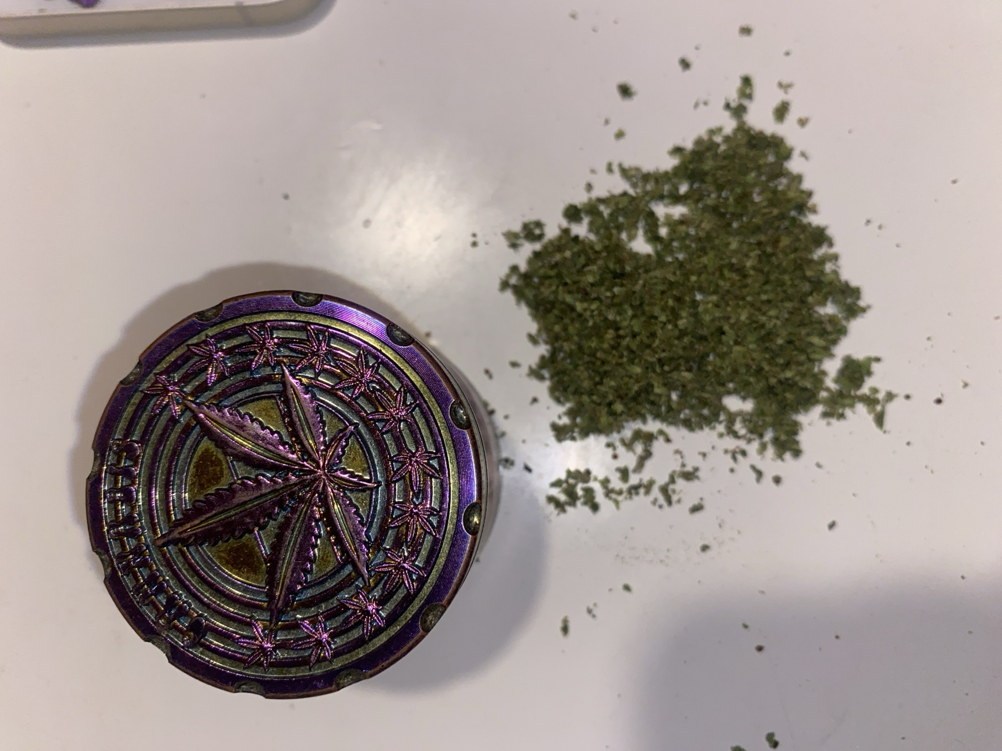 This image displays an overhead view of a metallic herb grinder and a quantity of ground green plant material on a white surface. The scene is located in Buenos Aires, Argentina. The grinder, positioned in the upper left section of the frame, is cylindrical with an iridescent finish exhibiting purple, green, and blue hues. Its lid features a prominent central cannabis leaf design, encircled by concentric rings and a border of smaller cannabis leaf motifs. The text "ORIGINAL" is discernible on the top rim of the grinder. To the lower right of the grinder, an irregular pile of finely ground, dark green botanical matter, consistent with dried cannabis, is present. Scattered smaller fragments of this material extend outwards from the main pile. The background is a uniform, smooth white surface, likely a table or countertop. No individuals or active interactions are visible.