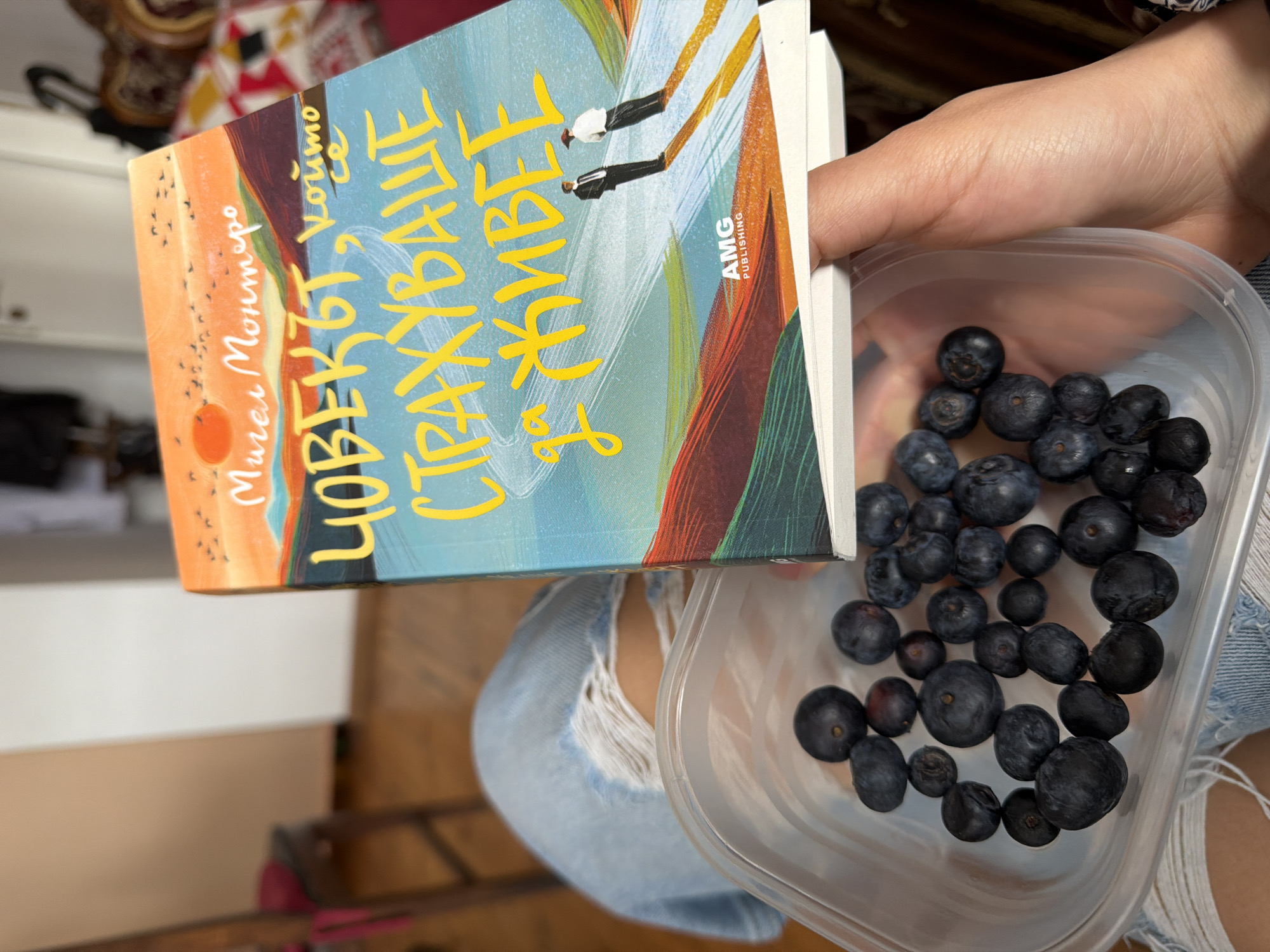 The image presents a downward-angled perspective on a person's lap and hands within an indoor residential environment, likely located in Sofia, Bulgaria. The subject's lower thighs are visible, covered by light blue ripped denim jeans. A clear plastic rectangular container, partially filled with approximately two dozen dark blue blueberries of various sizes, rests on the left thigh area. The subject's right hand grasps a paperback book titled "ЧОВЕКЪТ, който се СТРАХУВАШЕ да ЖИВЕЕ" (The Man Who Was Afraid to Live) by Miguel Montero, published by AMG Publishing. The book cover features a vibrant illustration depicting an orange sky with a sun and birds, rolling hills in shades of green and brown, a light blue body of water, and two small, stylized male figures standing on a path. The background, out of focus, reveals a light-colored wooden floor, a wooden chair with a pink cushion positioned to the left, and a patterned fabric object, possibly upholstery with red, white, and brown geometric designs, to the right. A light-colored wall and a partially visible white door are discernible further in the background.