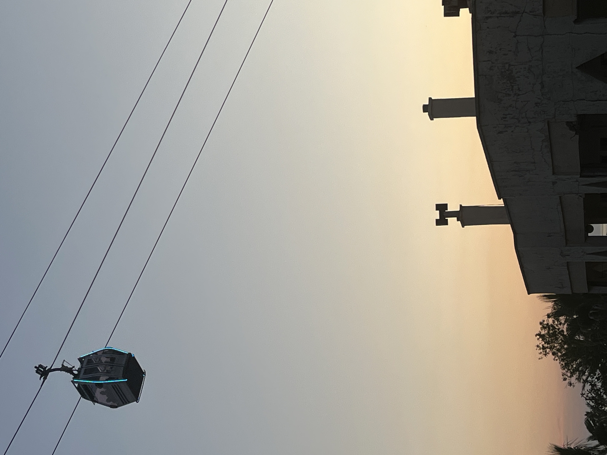Here's a description of the image:
The image, taken in what appears to be the late afternoon or early evening given the warm sunset tones in the sky, depicts a cable car in Alanya, Turkey. The cable car is of a modern design, possibly featuring some blue LED lighting around its edges. It is suspended from multiple cables that stretch diagonally across the left side of the image. To the right, the silhouette of a building structure with industrial-looking chimneys or vents stands out against the colorful sky. The very bottom right corner features glimpses of palm trees, a common element in the landscape of Alanya. The overall scene conveys a sense of travel and modern infrastructure integrated into a scenic environment.
