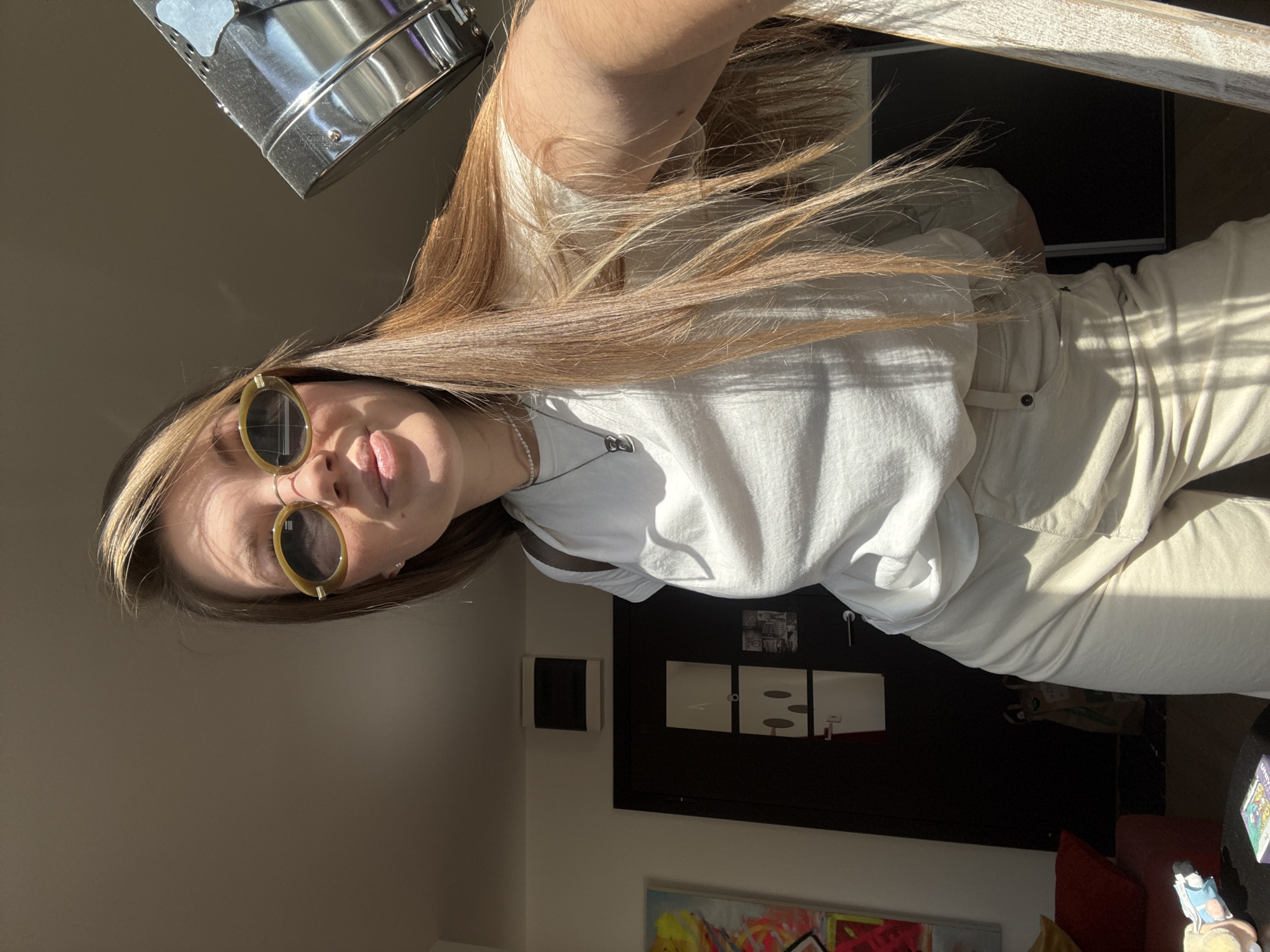 A young woman wearing sunglasses, a white t-shirt, and light-colored pants poses indoors in Vozdovac Urban Municipality, Serbia. She has long, light brown hair. A metal object is positioned at the top left corner of the image. Behind her is a wall with a black door and a small white air conditioning unit. A framed painting is visible near the bottom of the image. A table with various objects is in the bottom right corner. The scene is brightly lit with sunlight.