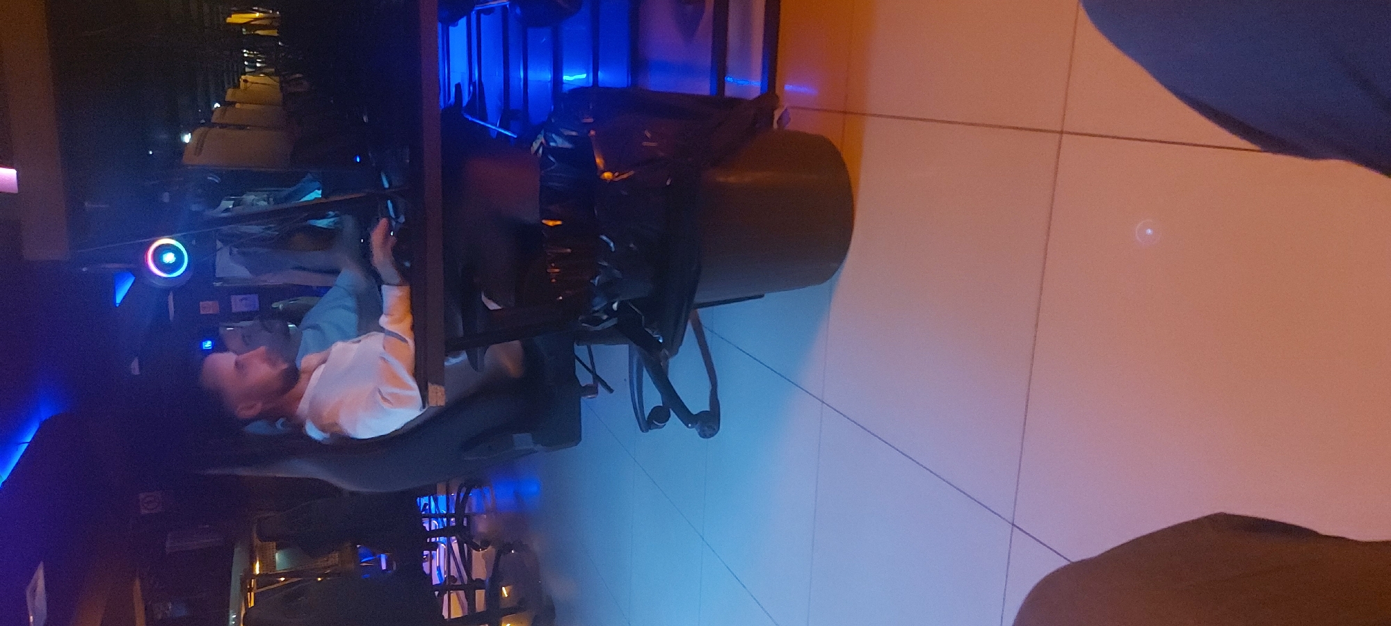 A man with a beard and a white shirt is seated at a desk with computer equipment in a brightly lit, modern room, likely in a gaming or co-working space. The location is plausibly in Tashkent, Uzbekistan, given the lack of easily identifiable cultural elements specific to other regions. Visible on the desk is a computer with LED lighting, a headset, and other accessories. The room has a tile floor and appears to be a public area, based on the presence of other people or furniture in the background.