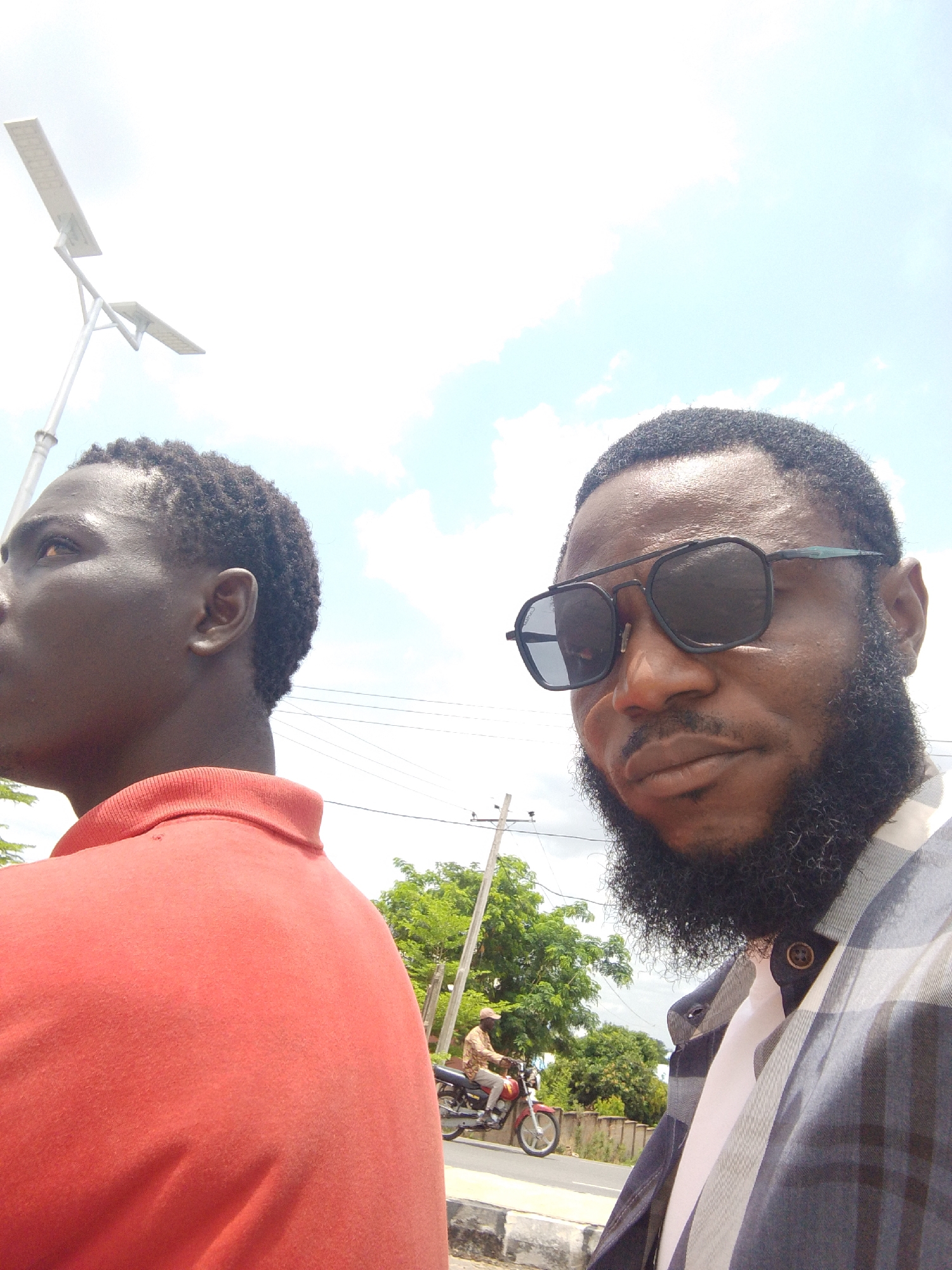 This is a brightly lit outdoor photograph featuring two men in the foreground and a street scene in the background. The image appears to be taken from a slightly low angle, looking upwards towards the sky.
On the right, a man with a dark, full beard and mustache is prominently featured, wearing dark-framed sunglasses. He has dark, short hair and is dressed in a plaid shirt with shades of blue/black and white, worn over a white collared shirt. He is looking directly towards the camera with a neutral expression.
To his left, partially obscured, is another man with dark, short, curly hair, seen in profile. He is wearing a red collared polo shirt and is looking towards the left side of the frame, away from the camera. His skin appears dark, reflecting the bright sunlight.
The background consists of a clear, bright sky with scattered white clouds. To the far left, a modern solar-powered street light on a tall pole is visible. Further back, towards the center and right, green trees line what appears to be a road or paved area. A utility pole stands among the trees. In the mid-ground, on the road, a person is seen riding a red motorcycle, moving from left to right. The general setting suggests a roadside or urban fringe environment in Lokoja, Nigeria, consistent with the visible infrastructure and vegetation.
The time of day is clearly daytime, likely midday or early afternoon, given the intensity of the light and the high position of the sun. There is no visible text in the image. The overall mood is casual and observational.
