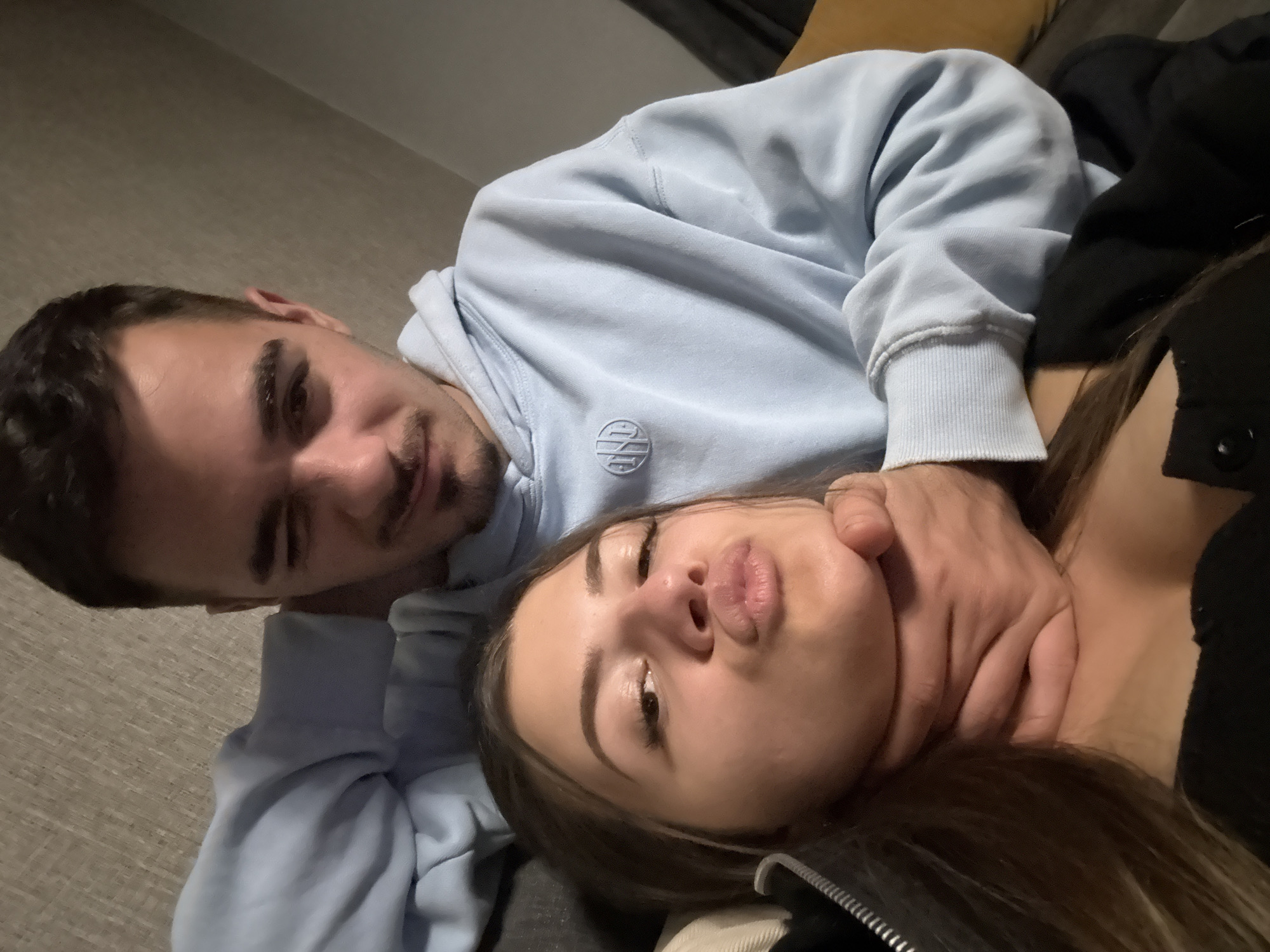 The image, captured in an indoor setting in Sofia, Bulgaria, features two individuals, a man and a woman, in a close-up, tilted shot that appears to be a selfie or intimate snapshot.
The man, positioned in the background and to the left, has short dark hair, a beard, and dark eyebrows. He is wearing a light blue hooded sweatshirt that features a circular emblem with a stylized design (possibly intertwined letters) on the chest. He gazes towards the viewer with a slight smirk or playful expression.
In the foreground and to the right, the woman has long dark hair and is dressed in a dark top. Her head is tilted, and she is making a "pouty" or "kissing" face with slightly parted lips, indicating a lighthearted and affectionate mood.
A central element of their interaction is the man's right hand, which is gently placed around the woman's neck. This gesture, combined with their expressions, conveys a sense of playful intimacy rather than aggression. The background shows a neutral-toned, possibly carpeted floor or wall on the left, and a darker upholstered surface on the right, suggesting a casual home environment, possibly a sofa or bed. The lighting is artificial, common for indoor nighttime or subdued daytime settings. The overall mood of the image is one of tender playfulness and closeness.