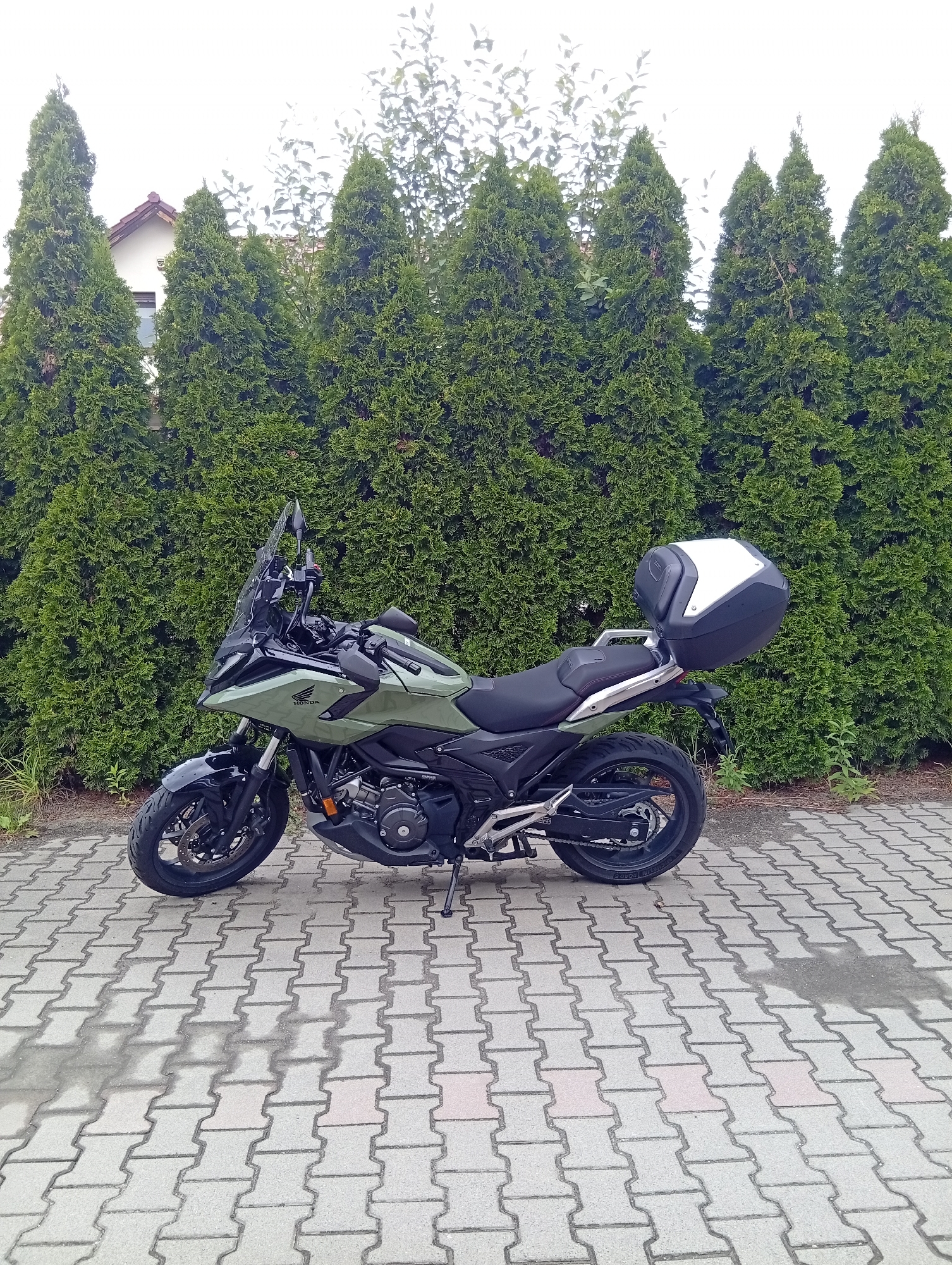 A green Honda motorcycle with a black and white top case is parked on a paved driveway in front of a dense line of green evergreen trees. A portion of a white building with a red roof is visible behind the trees on the left side of the image. The motorcycle is angled slightly towards the right. The pavement is made of interlocking gray pavers. The sky is overcast and white, with some faint greenery visible at the top of the image. There are no people present in the image. The scene is set outdoors, likely in a residential area.