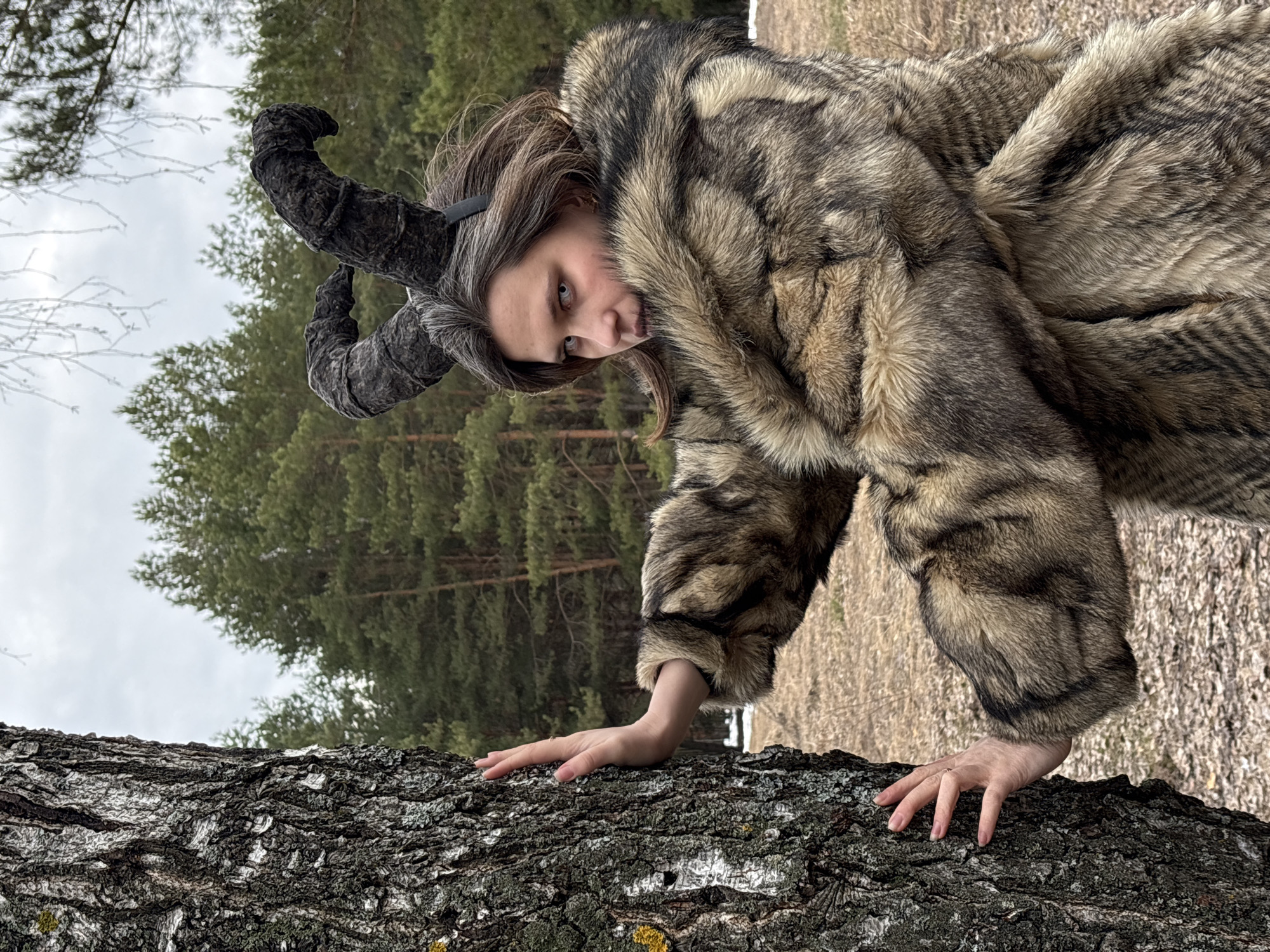 A person dressed in a wolf-like fur coat with large, curved horns is positioned in front of a tree trunk. The person is bending forward, placing their hands on the rough surface of the tree. The background features a wooded area with evergreen trees, set against an overcast sky. The location is potentially Chaikovsky, Russia.
