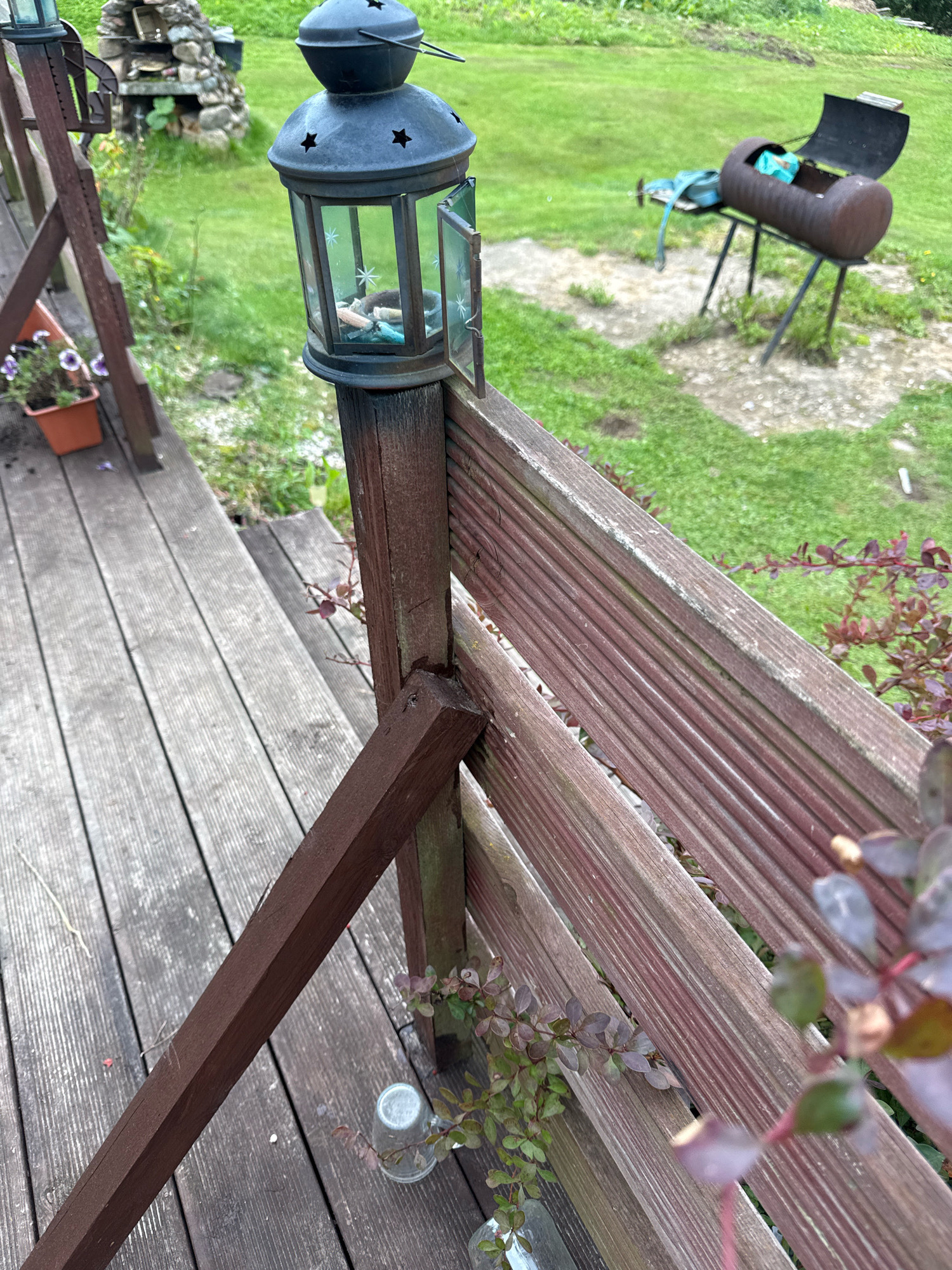 The image depicts a weathered wooden deck and railing with a vintage lantern mounted on a wooden post. The lantern is made of dark metal with glass panes and star cutouts. Inside the lantern, small decorative items are visible. In the background, a green grassy area is present, with a rustic grill and a small stone structure. The scene appears to be outdoors, possibly in a backyard or garden setting, with hints of a rural environment. The location of Galinė, Lithuania is not depicted or indicated in the image.