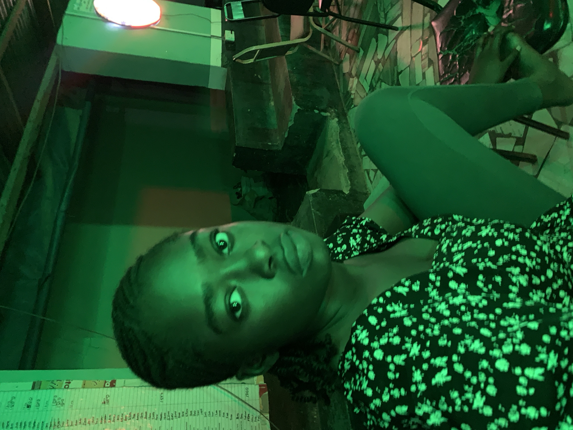 An eye-level, tilted portrait captures a young woman in a dimly lit indoor setting, heavily saturated with green light. The woman, appearing to be of African descent with dark skin and braided hair, is positioned diagonally across the frame. She looks directly at the camera with wide, dark eyes, maintaining a neutral to slightly curious expression. She is wearing a dark top adorned with a light, speckled or floral pattern that glows under the green illumination, paired with dark leggings or trousers.
The background features indistinct dark furniture, possibly a chair or stool and a table, along with a textured or tiled wall behind it. A prominent, bright, reddish-orange circular light source is visible in the upper part of the frame, contributing to the artificial lighting scheme. Below the woman's head, a white sheet of paper is visible, covered in columns of faint text and numbers, suggesting a menu, price list, or an inventory. Numbers such as "1500," "100," "500," and "200" are faintly discernible amidst other illegible script.
The dominant green light creates a distinctive, moody, and artificial atmosphere, indicating a nighttime setting or an indoor venue like a bar, lounge, or restaurant. The casual posing suggests a personal photo or selfie taken in New Stadium, Nigeria.