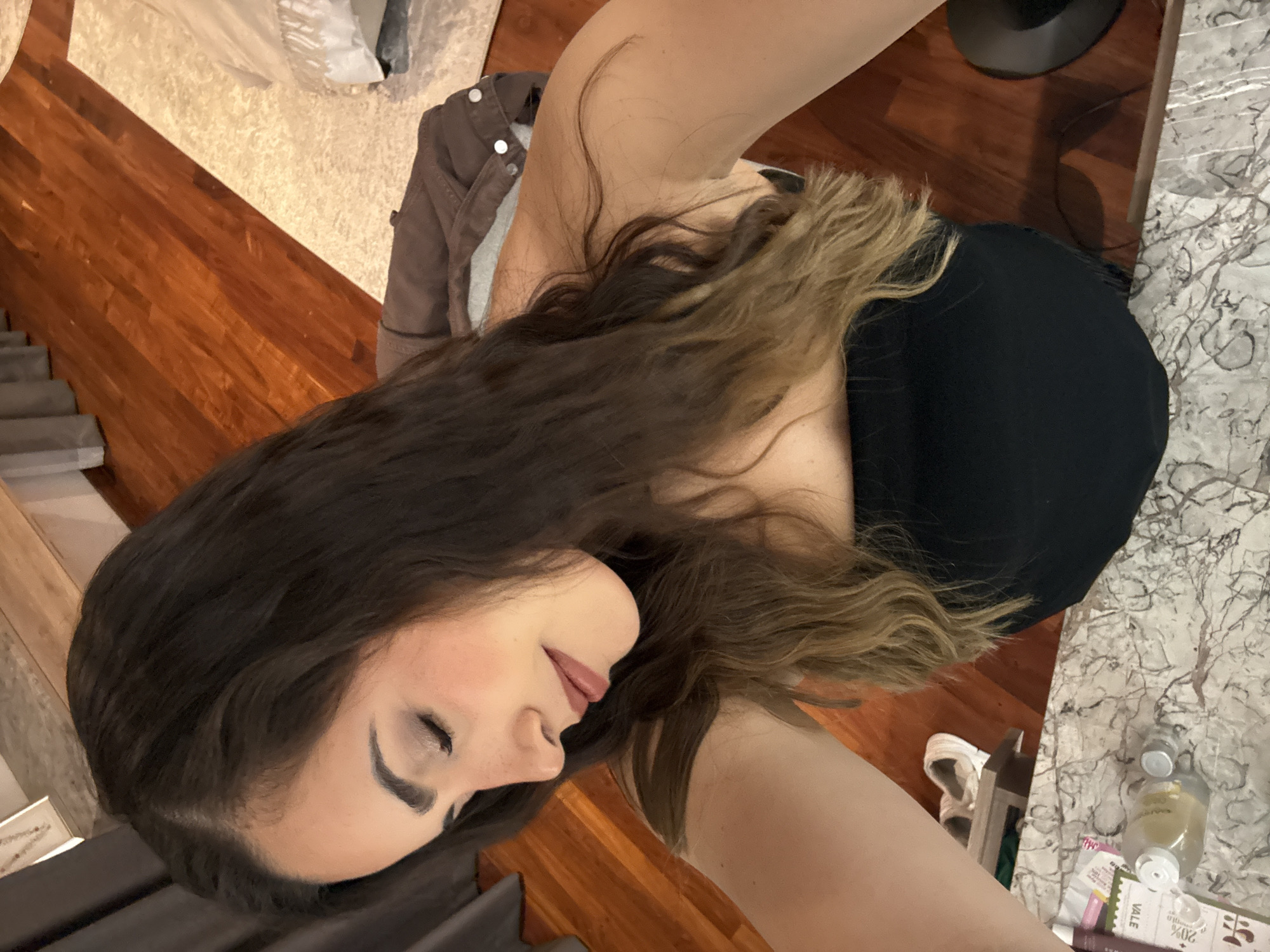 A woman with long brown hair and fair skin is seen in a close-up, high-angle shot. She is wearing a black top and a jacket. The background features a mix of interior elements, including wood flooring, a textured rug, and a marble countertop. There are also various items on the countertop, such as bottles and cosmetic products. The photo appears to be taken indoors in what seems to be a residential setting. The location in San Bernardino Tlaxcalancingo, Mexico cannot be verified based on the image alone.