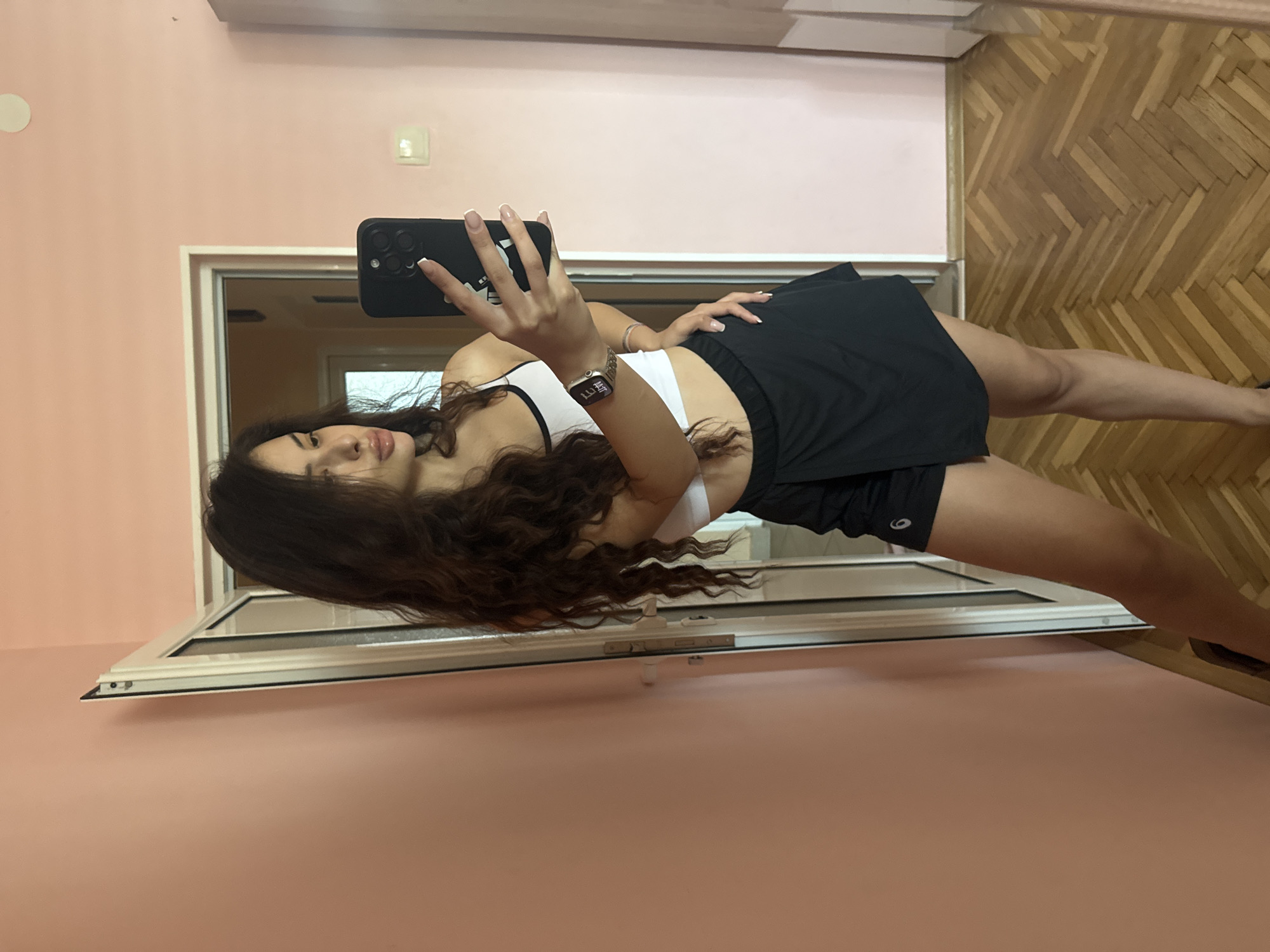 A young woman with long, dark, curly hair is taking a mirror selfie in what appears to be a bedroom. She is wearing a white crop top with black trim and a black skirt that has a skirt-like overlay. On her left wrist, she has a smartwatch. She is holding a black smartphone in her right hand to take the photo. The room has pale pink walls and a wooden floor laid in a herringbone pattern. To the left of the woman is a white door frame, open, revealing a glimpse of another room. The overall lighting suggests it is daytime. The image does not contain any elements that would allow for a specific location within the city of Plovdiv, Bulgaria to be identified.