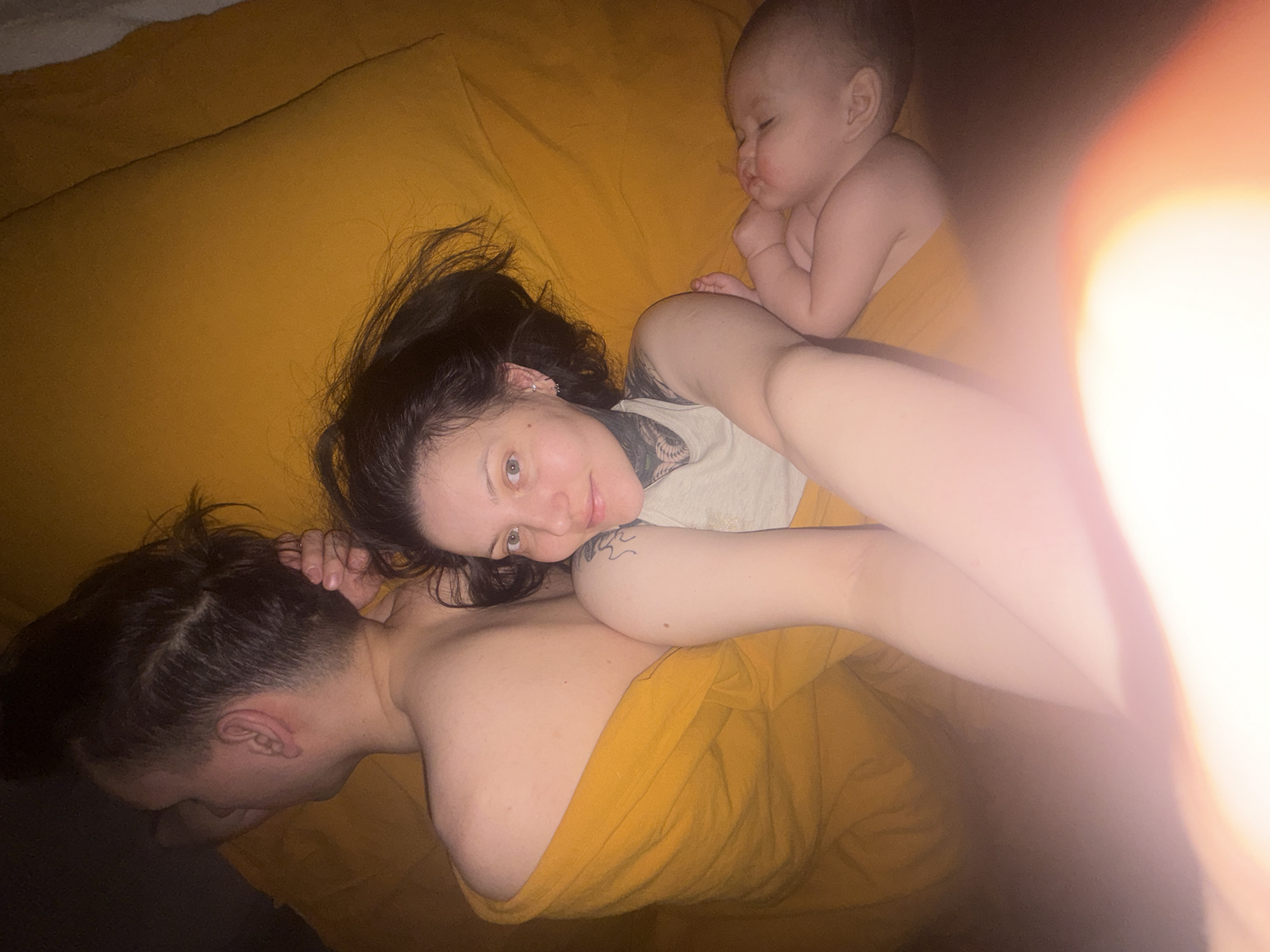 A photograph, possibly taken indoors in a private residence, features three individuals. At the bottom left of the frame, a person with short, dark hair is lying on their stomach, facing away from the camera. Above them and slightly to the right, a woman with long, dark hair and tattoos is lying on her side, looking towards the camera. A baby is positioned above the woman and to the right, also lying down. The scene is illuminated by warm, possibly artificial light, with an area of bright light obscuring the right side of the frame. All three individuals are lying on a mustard-yellow-colored surface, possibly a bed or a large cushion. The specific location in the city of Chaikovsky, Russia, cannot be determined from the visual information provided.