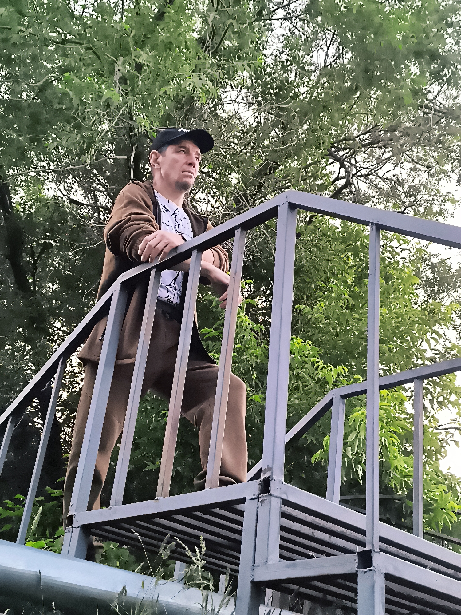 A man in a brown tracksuit and a black baseball cap stands on a metal staircase, leaning on the railing. He is looking to the right with a pensive expression. The staircase is made of grey metal with horizontal bars for steps and vertical supports. Lush green foliage from trees forms the background, with some sunlight filtering through the leaves. The scene appears to be outdoors, possibly in a park or an industrial area. The image does not provide any specific visual cues to pinpoint the location within the city administration of Qarağandı, Kazakhstan.