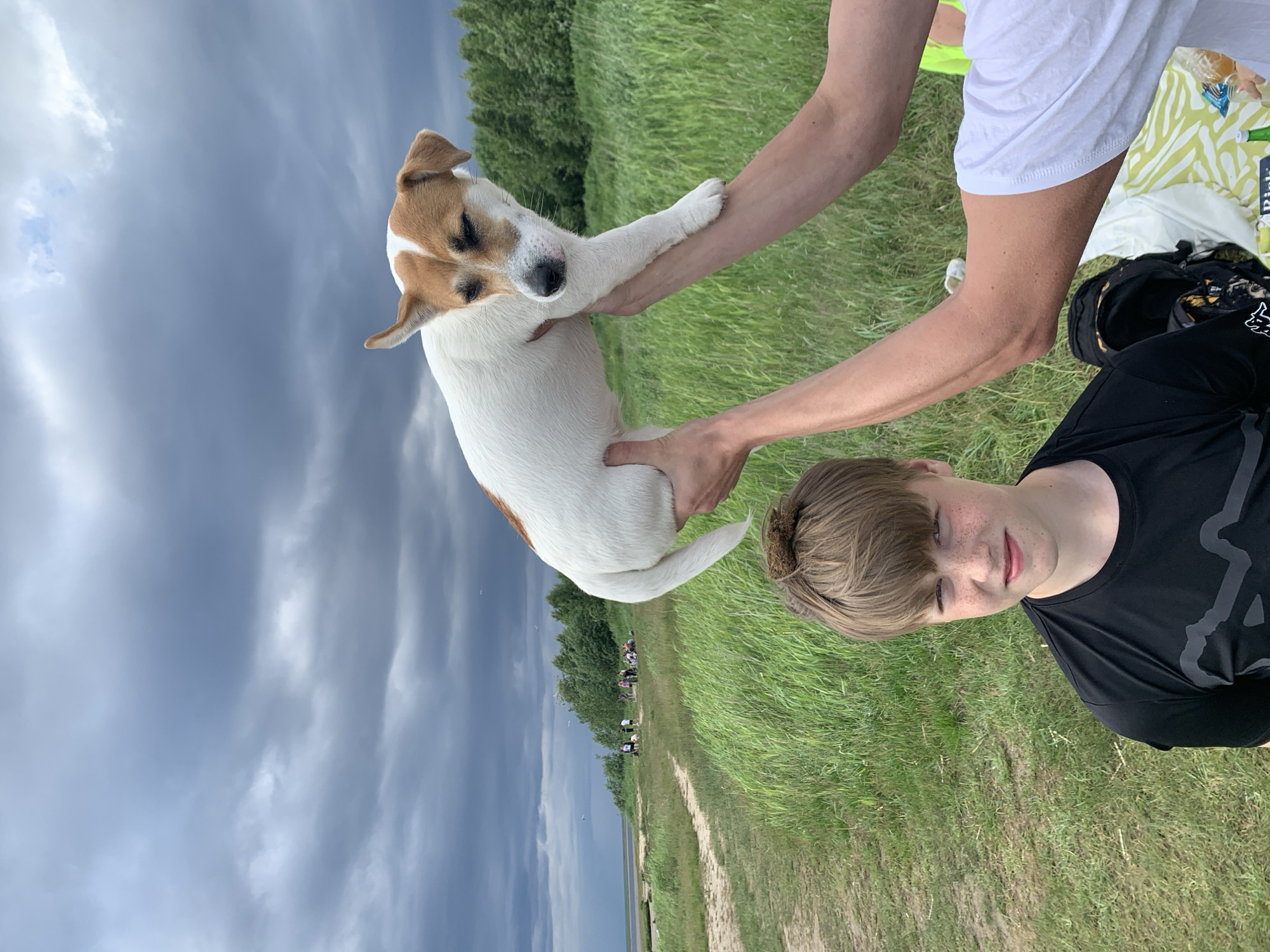 In a grassy field with a cloudy sky overhead, two individuals are interacting with a small dog. The dog, a Jack Russell Terrier, is being held by one person, its body white with brown markings on its head and back. Its eyes are closed, and it appears to be relaxed. The person holding the dog has their arms around its body, supporting it.
In the foreground, the face of a young person with light brown hair and freckles is visible. They are looking slightly to the side with a neutral expression. They are wearing a black t-shirt with a grey graphic logo on it. Behind them, another person is partially visible, wearing a white t-shirt and with their arms around the dog.
In the background, a path winds through the field, leading towards a line of trees. Beyond the trees, a body of water can be seen, and further in the distance, a few small figures are present. The overall scene suggests an outdoor gathering or outing, possibly a picnic or a walk in a park-like setting. The time of day appears to be daytime, with the cloudy weather contributing to a somewhat diffused light. There is no visible text in the image.