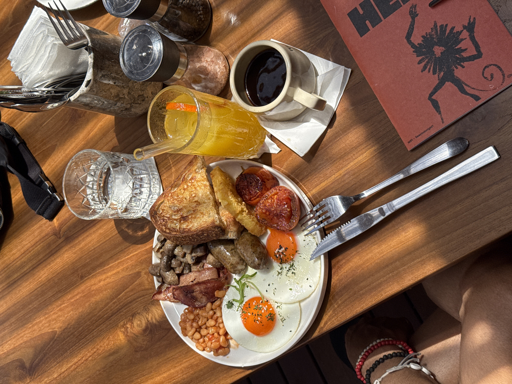 On a wooden table, an overhead shot displays an English breakfast with fried eggs, sausage, bacon, beans, tomatoes, hash browns, toast, and mushrooms on a white plate, accompanied by cutlery. A glass of orange juice with a straw and a coffee cup sit beside the plate, along with a glass of water, salt and pepper shakers, and a napkin holder filled with napkins and cutlery. A red menu with a graphic of a dancing figure is also present. An individual's leg, adorned with bracelets, is visible in the bottom right corner. The scene suggests a casual dining experience in Kuta Selatan, Indonesia, though the specific location within the city cannot be determined from the image alone.