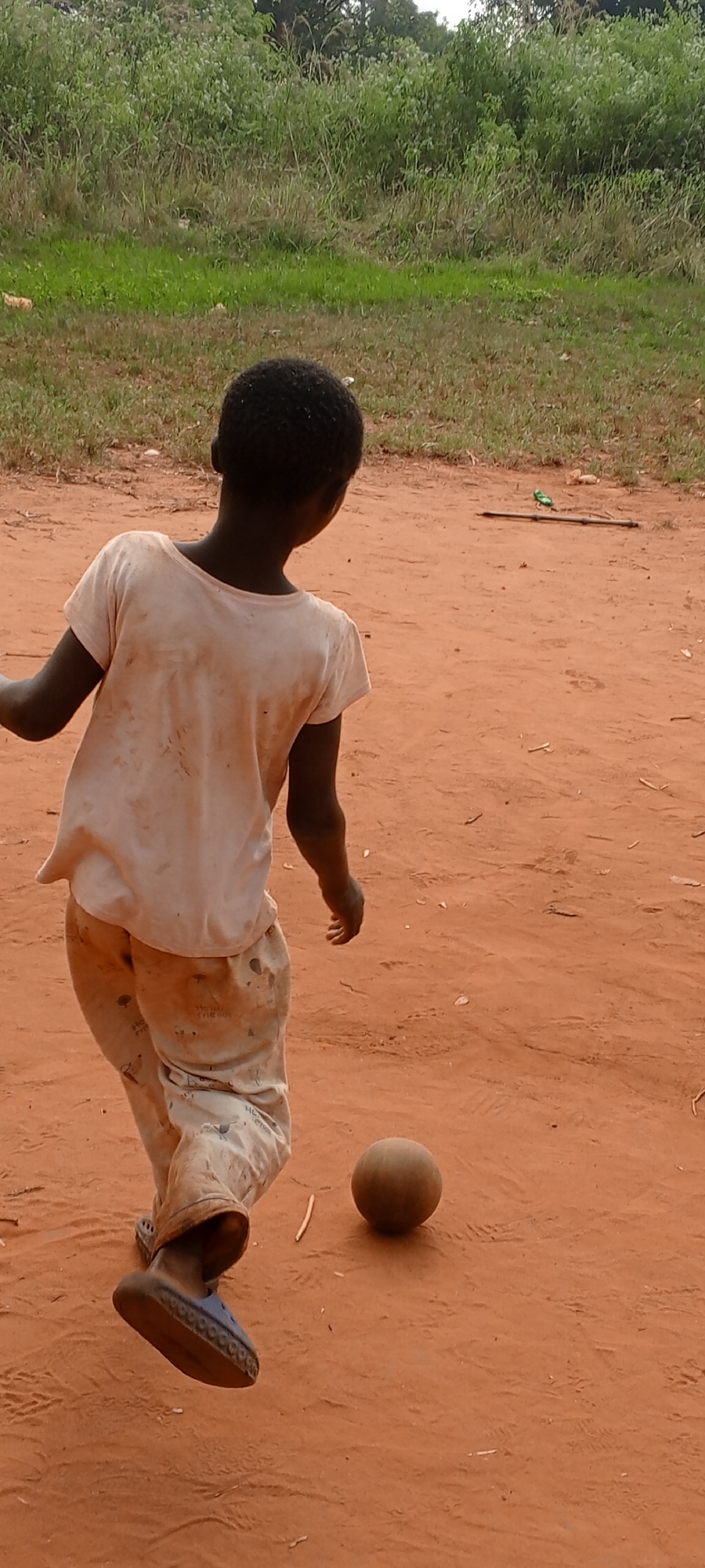 A young Black child stands on red-brown, dusty ground in an outdoor setting. The child wears a dirty, light-colored t-shirt and paint-splattered pants, with blue sandals on their feet. The child’s back is to the viewer, and they appear to be looking at a brown ball in front of them. Green vegetation is visible in the background. A stick lies on the ground in the distance. The photo was taken in Ngalula, Democratic Republic of the Congo.