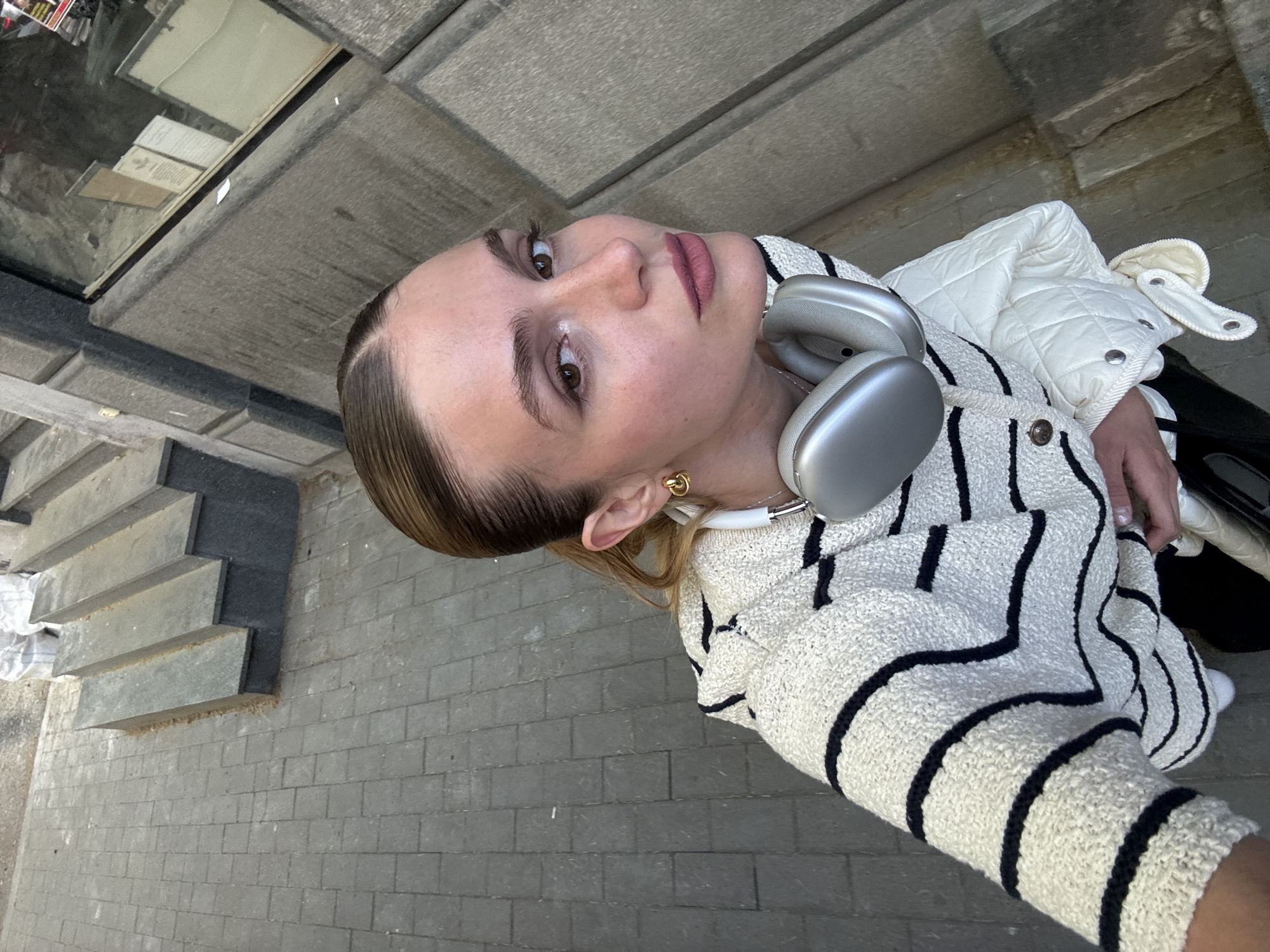 A woman is taking a selfie in Tbilisi, Georgia. She is standing on a gray-paved street next to a staircase that leads to a building with a glass facade reflecting the street. The woman is wearing a cream-colored jacket with thick black stripes and a white quilted jacket over it. She has headphones around her neck and is wearing gold earrings. The photo is taken at an angle, showing the street and building behind her.