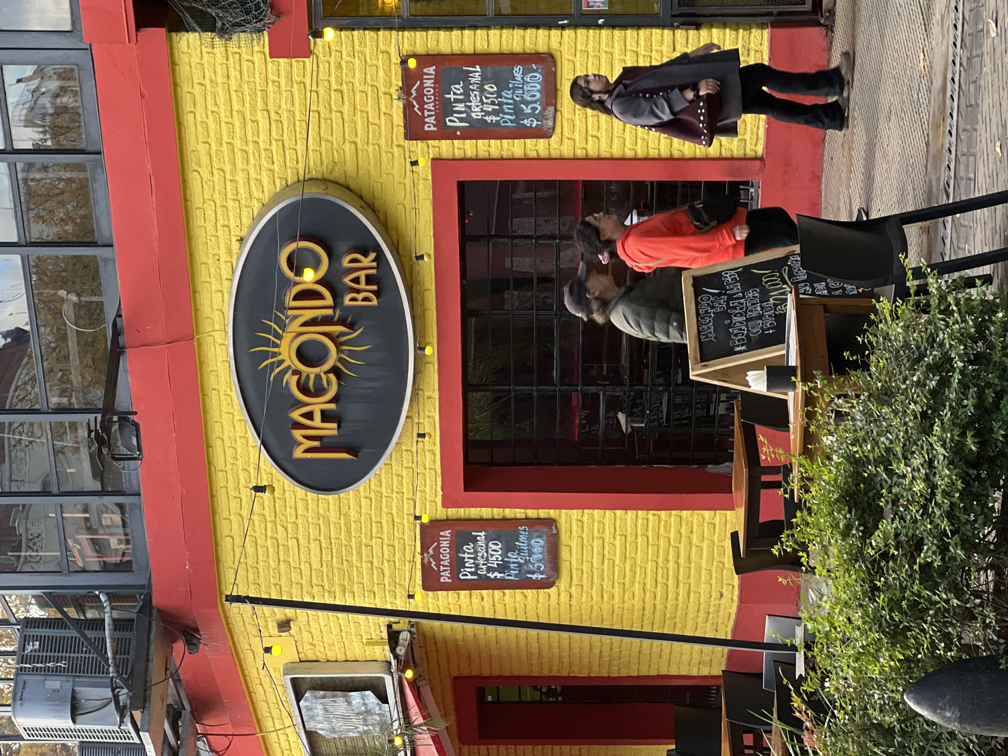 The image shows the exterior of "Macondo Bar" in Buenos Aires, Argentina. The building has a distinctive yellow brick facade with red trim around windows and edges. A large, oval-shaped sign with "MACONDO BAR" is prominently displayed, featuring a sun-like image beneath the name. Two chalkboards are visible, one advertising "Pinta artesanal" and "Pinta Quilmes" with prices, and another promoting "Bondiolla Libre con Fritas". Three people are partially visible near the entrance. The foreground includes outdoor seating with chairs and a table partially obscured by a green bush.