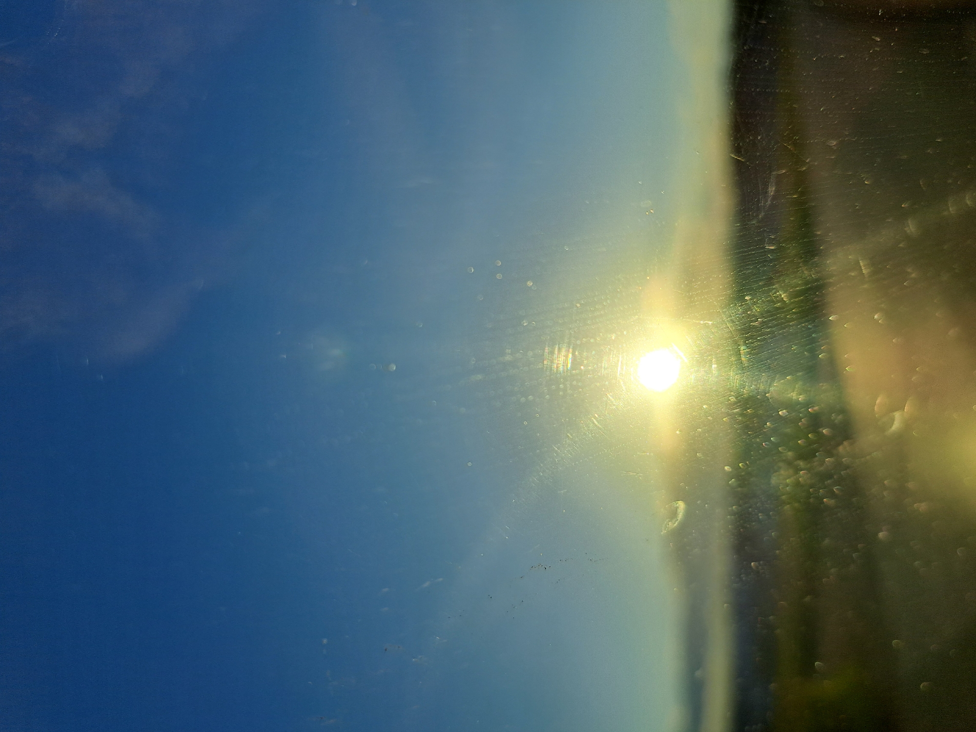 The image shows a bright sun low in the sky, casting a warm glow. The sun appears to be shining through a dirty or streaked window, creating lens flares and distorting the view of the landscape below. The landscape itself is out of focus, appearing as a blurred band of greens and browns, suggesting it is likely a natural or rural setting rather than an urban one. The sky is a clear blue, with a few wispy clouds. There are no people, animals, or specific man-made structures clearly visible. Given the lack of discernible urban elements, it is not possible to determine if this scene is located in the city of Chernetsk, Russia. The image composition and focus suggest it might be a view from a vehicle or a window in a rural or suburban area.