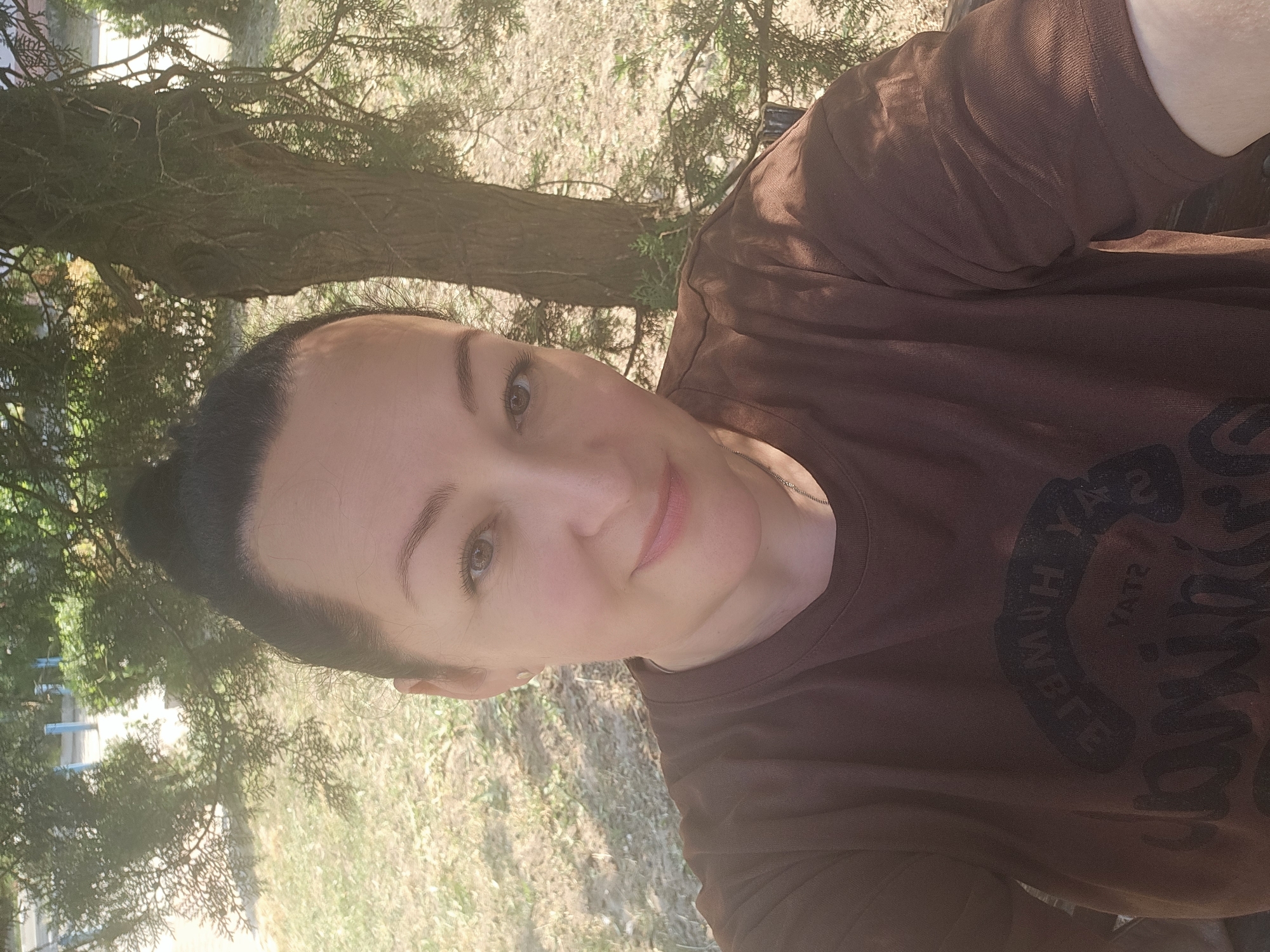 A woman is pictured outdoors, possibly during the day. She has dark hair pulled back and is wearing a brown t-shirt with dark lettering. Her expression is neutral. She appears to be in a park or natural setting with trees and dry grass. The lighting suggests it is daytime. There are some blue structures visible in the background, which could be park benches or playground equipment. The image seems to be a selfie, taken from a slightly low angle. There is some text on the t-shirt, but it is difficult to read clearly due to the image quality and angle.