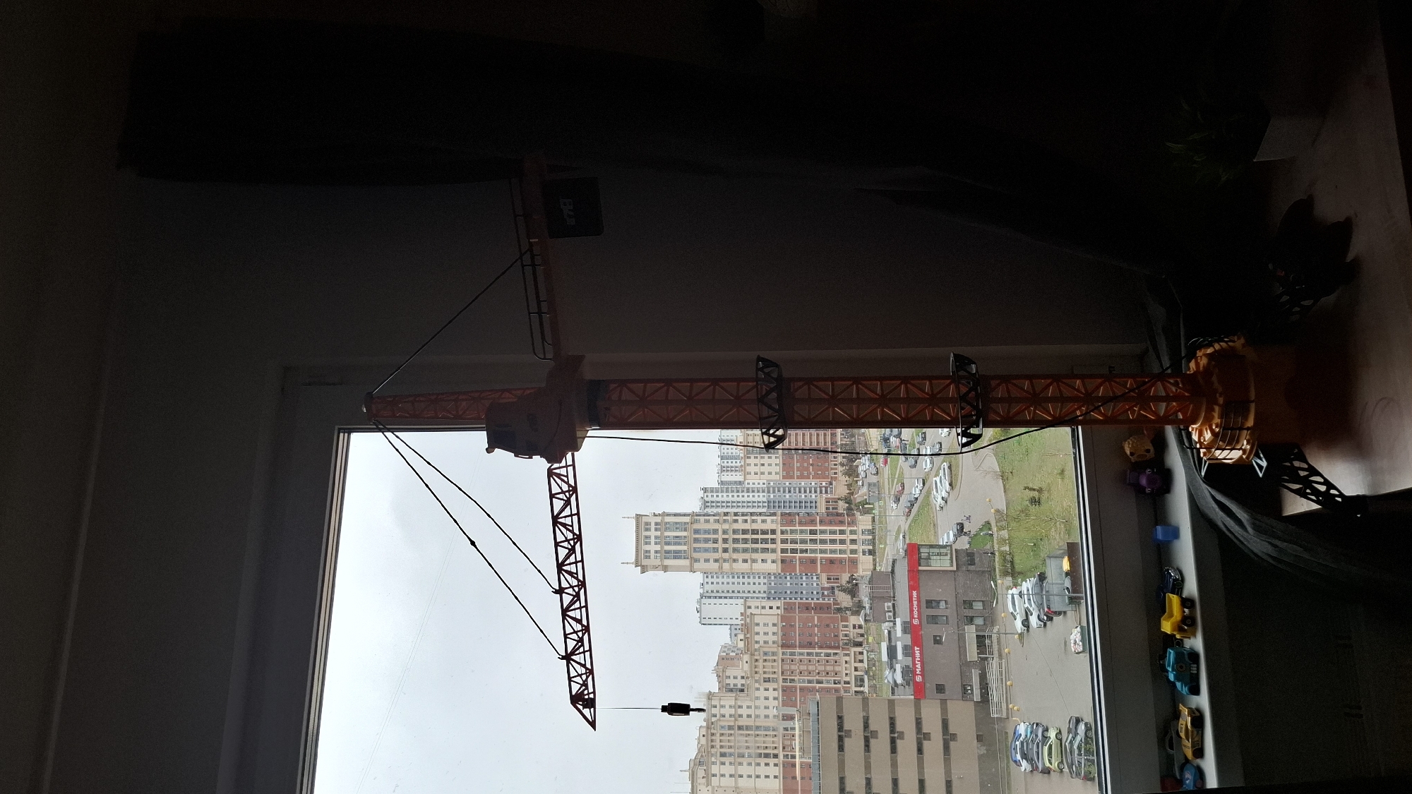 The image captures an indoor view looking out a window, dominated by a large, orange and black model crane positioned on the windowsill. The crane's main boom extends across the upper part of the window frame, with its counterweight section on the right (as if upright) and the jib extending leftwards. Several small toy cars (including yellow, blue, and black vehicles) and miniature figures (such as a purple character and a yellowish blob) are arranged on the windowsill next to the crane's base. The interior space to the left and top of the window appears dark, suggesting low light conditions indoors.
Through the window, an urban landscape is visible under an overcast sky. Multiple multi-story apartment buildings, varying in height and color (including beige, white, and red-brick facades), form the backdrop. Below the buildings, a parking lot filled with numerous cars is discernible, alongside patches of green lawn.
Notable details include a red commercial sign on a building in the mid-ground, which clearly displays the Russian text "МАГНИТ" (Magnit) and "РОСНЕФТЬ" (Rosneft), consistent with the provided Saint Petersburg, Russia location context. The sky is uniformly grey and cloudy, indicating an overcast day with no direct sunlight. No human activity is depicted in the scene; the setting is static, featuring toys by a window overlooking a city street. The overall impression is of a quiet, somewhat dim daytime scene.