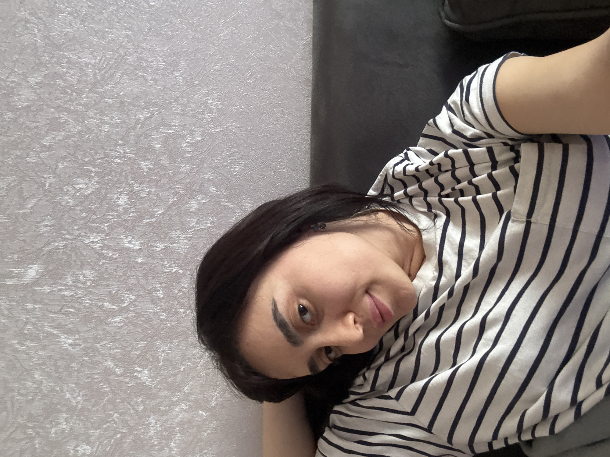 A young woman with dark hair is pictured from a tilted angle, wearing a striped shirt. She is lying against a light-colored, textured wall, with a dark gray surface visible near the top right corner. The location in Tbilisi, Georgia cannot be determined from the image.