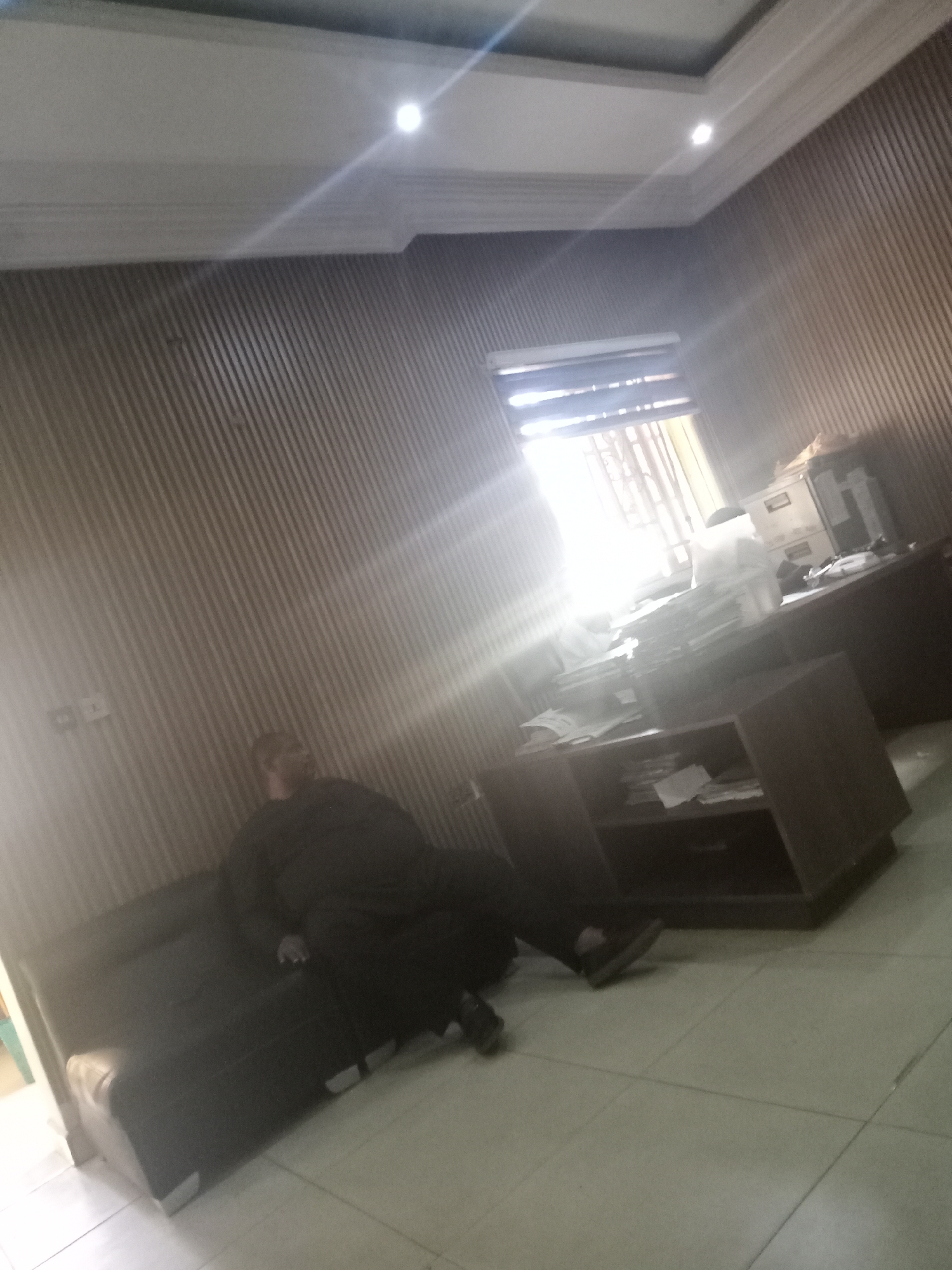 A person is reclined on a black couch in an office. The person is dressed in black attire. The office has wooden walls and tile flooring. A desk with papers and a filing cabinet are visible, suggesting the office is in use. The window is brightly lit. This is likely a workspace in Gwarinpa, Nigeria.