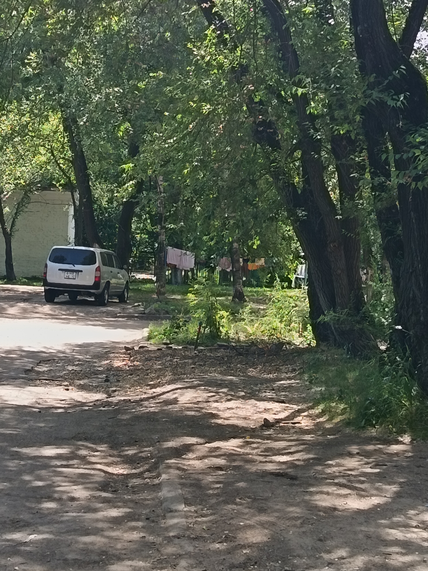 The image depicts a sun-dappled dirt road in Komsomolsk-on-Amur, Russia, with lush green trees lining the sides. In the background, a white van is parked on the left side of the road, facing away from the camera. Clothes are hanging on a clothesline to the right of the van. A small white building is partially visible behind the van. The ground is covered with dirt, gravel, and scattered leaves, with shadows cast by the trees creating a patterned effect. The overall scene suggests a residential area or a natural setting within the city, likely during a warm day.