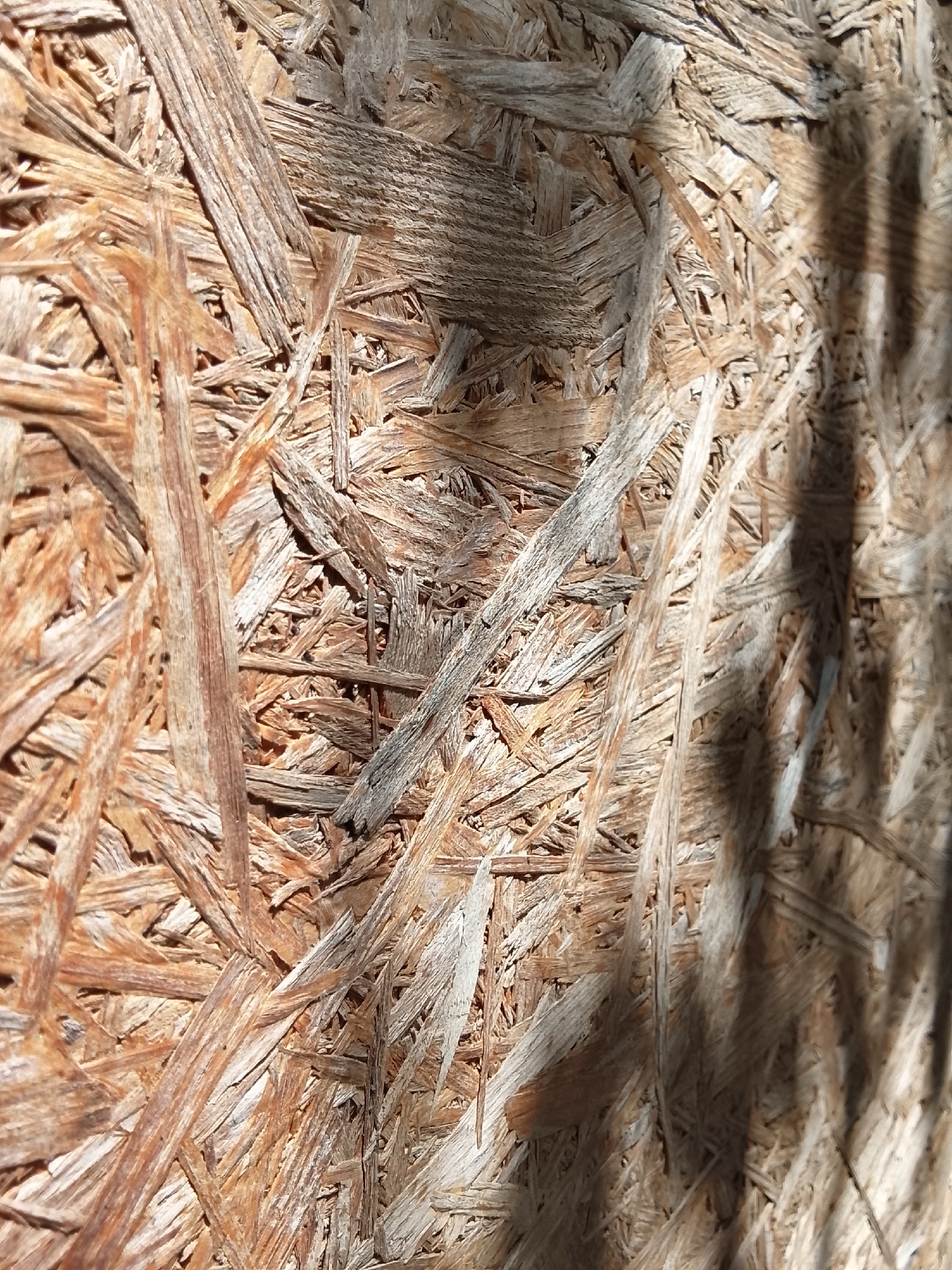 A close-up, full frame, eye-level shot focuses on a textured surface composed of compressed wood chips, creating a rough, uneven pattern. The material appears to be Oriented Strand Board (OSB) or a similar composite wood product. The wood chips are varied in size and shape, with dominant brown and beige tones, some exhibiting lighter, aged wood grains and darker, more resinous areas. The surface is lit by directional sunlight, casting distinct shadows across the texture, highlighting the three-dimensional nature of the material. The shadows, particularly visible on the right side of the frame, suggest an angled light source, possibly from a window or an outdoor structure. There are no people, animals, or discernible man-made objects other than the material itself. The overall impression is one of a weathered, natural building material. The location in the city of Жалпақсай, Kazakhstan, is not visually evident from the provided image.