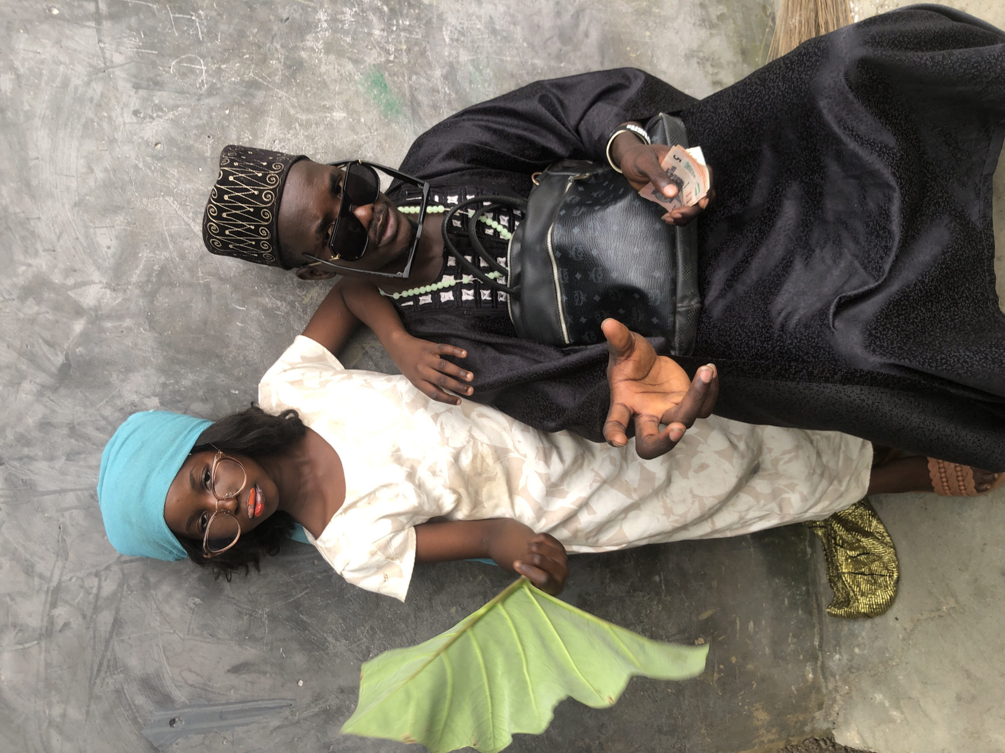 In Ado, Nigeria, two individuals pose against a textured grey wall. On the left, a young person wearing a bright teal headscarf and a cream-colored dress with a subtle floral pattern holds a large, vibrant green leaf. They are adorned with oversized, clear-rimmed glasses and have striking orange and red lipstick. To the right, an adult, possibly male, wears a black kaftan with intricate white patterns, a black cap with gold detailing, and large black sunglasses perched on their nose. They are accessorized with a light green beaded necklace and a black MCM-patterned bag. The adult is also holding a small wad of cash in their outstretched hand, and a gold clutch rests on the ground near the feet of the younger person. The overall impression is one of posed and stylistic presentation in an urban outdoor setting.