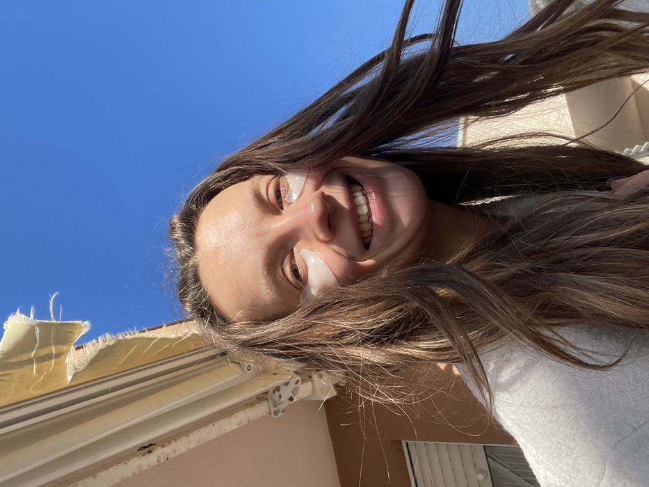 An adult female subject, with long brown hair, is depicted smiling directly at the camera in a close-up, outdoor portrait. She has white, translucent eye patches applied beneath each eye. The subject is illuminated by direct sunlight, which highlights her hair and face. She wears a light-colored, possibly white or cream-colored, fuzzy garment.
In the upper left background, a light-colored, worn fabric awning, featuring frayed edges, is visible, partially extended. Below the awning, a section of a building facade, light pinkish-beige in color, is present, with a white rolling shutter partially visible on the far left. White metal brackets securing the awning are also visible. The upper right and central background consist of a clear, bright blue sky.
The scene is set outdoors, likely on a balcony or near a window of a residential building, in the city of Budva, Montenegro, under clear weather conditions.