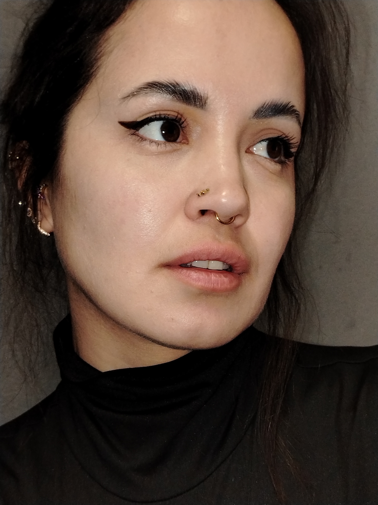 A close-up portrait of a woman with dark hair pulled back, wearing a black turtleneck shirt. She has fair skin, brown eyes, and distinct eyebrows. Her makeup includes eyeliner and lip gloss. She has multiple piercings, including a nose ring and studs, as well as earrings. The background appears to be a plain gray wall. The location is not identifiable as being in Noviy, Russia.